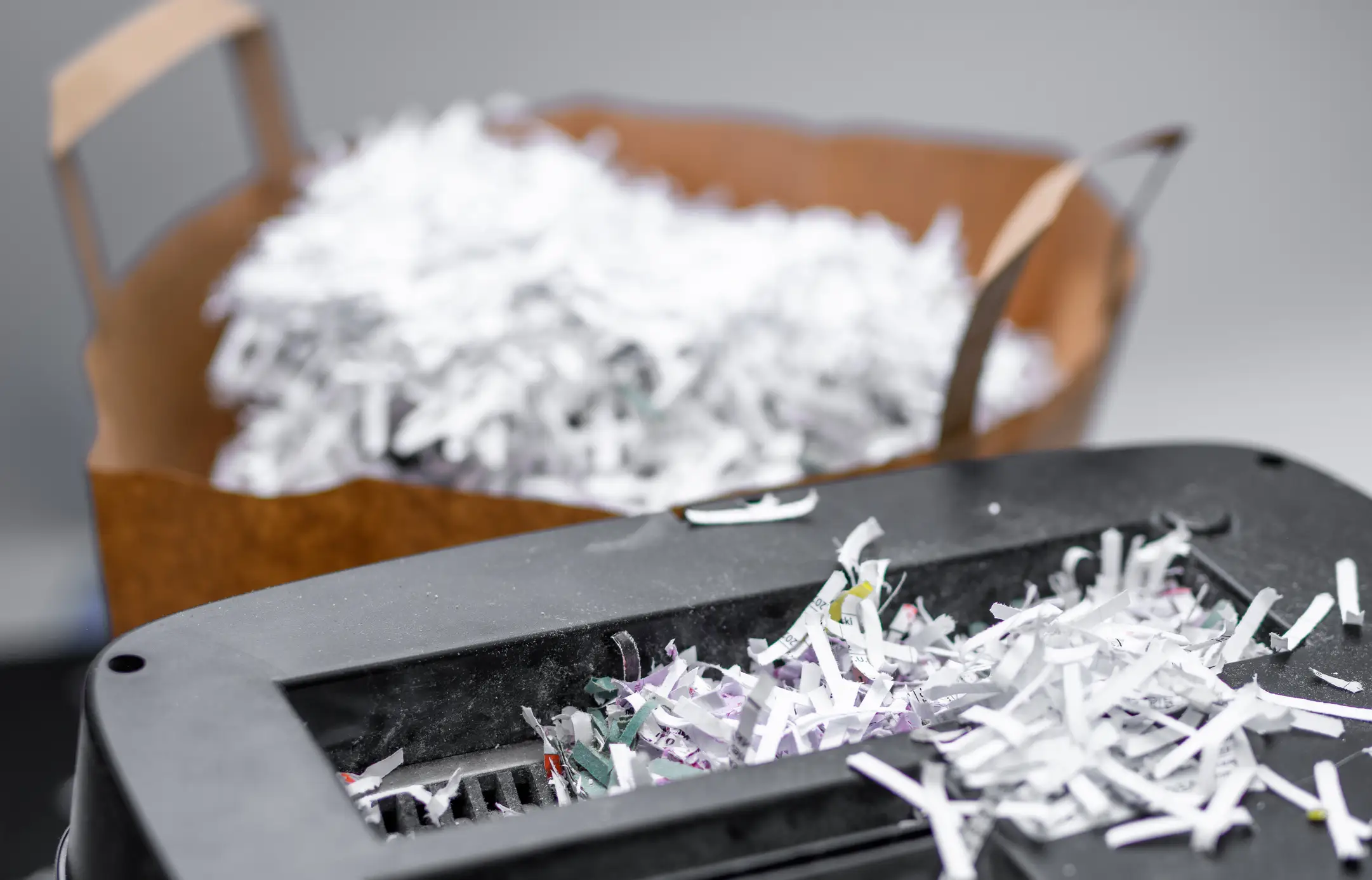 Paper shredder filled with shredded documents, with a basket of paper strips in the background.