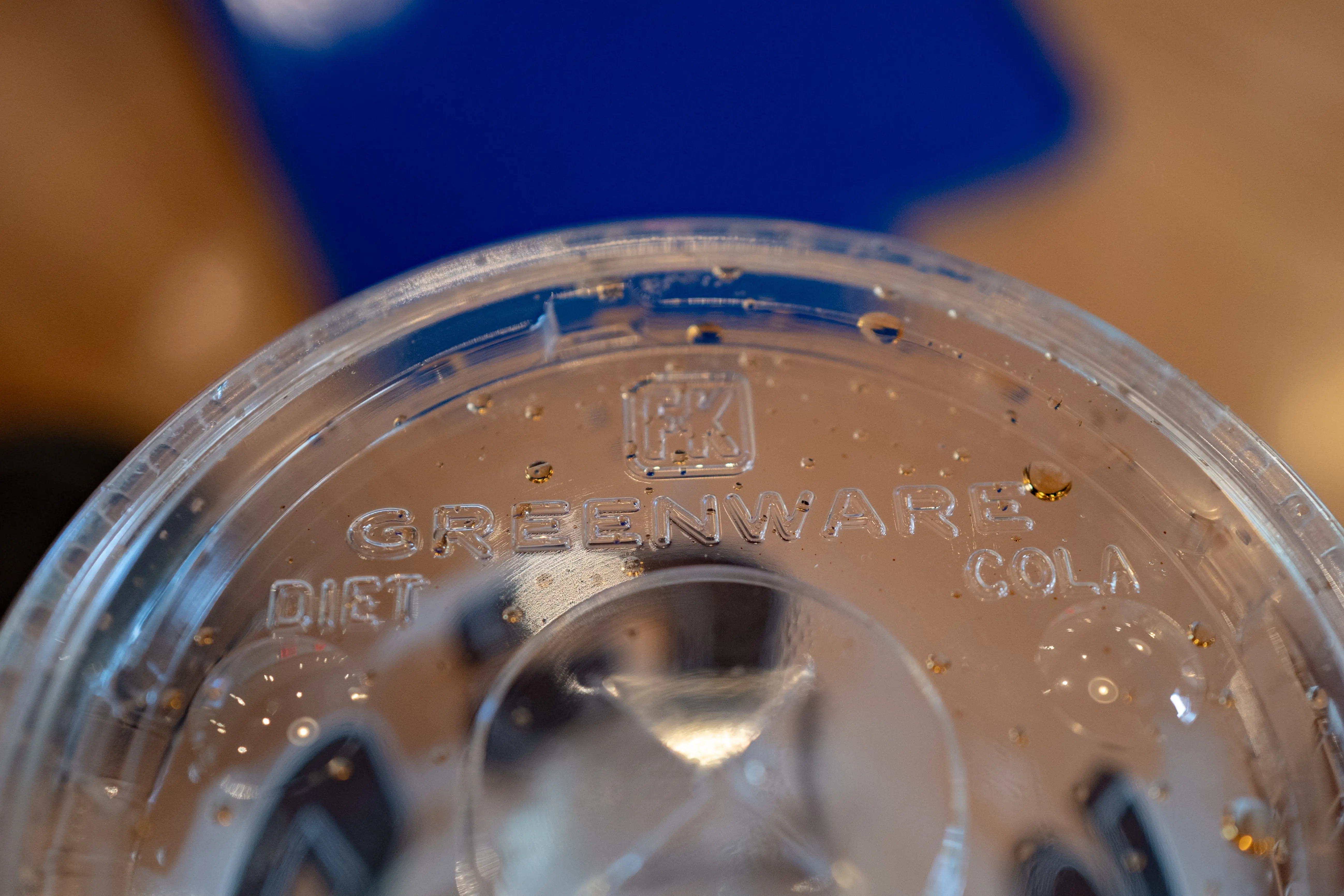 Close-up of a clear plastic drink lid embossed with GREENWARE, DIET, and COLA beside ice and droplets.