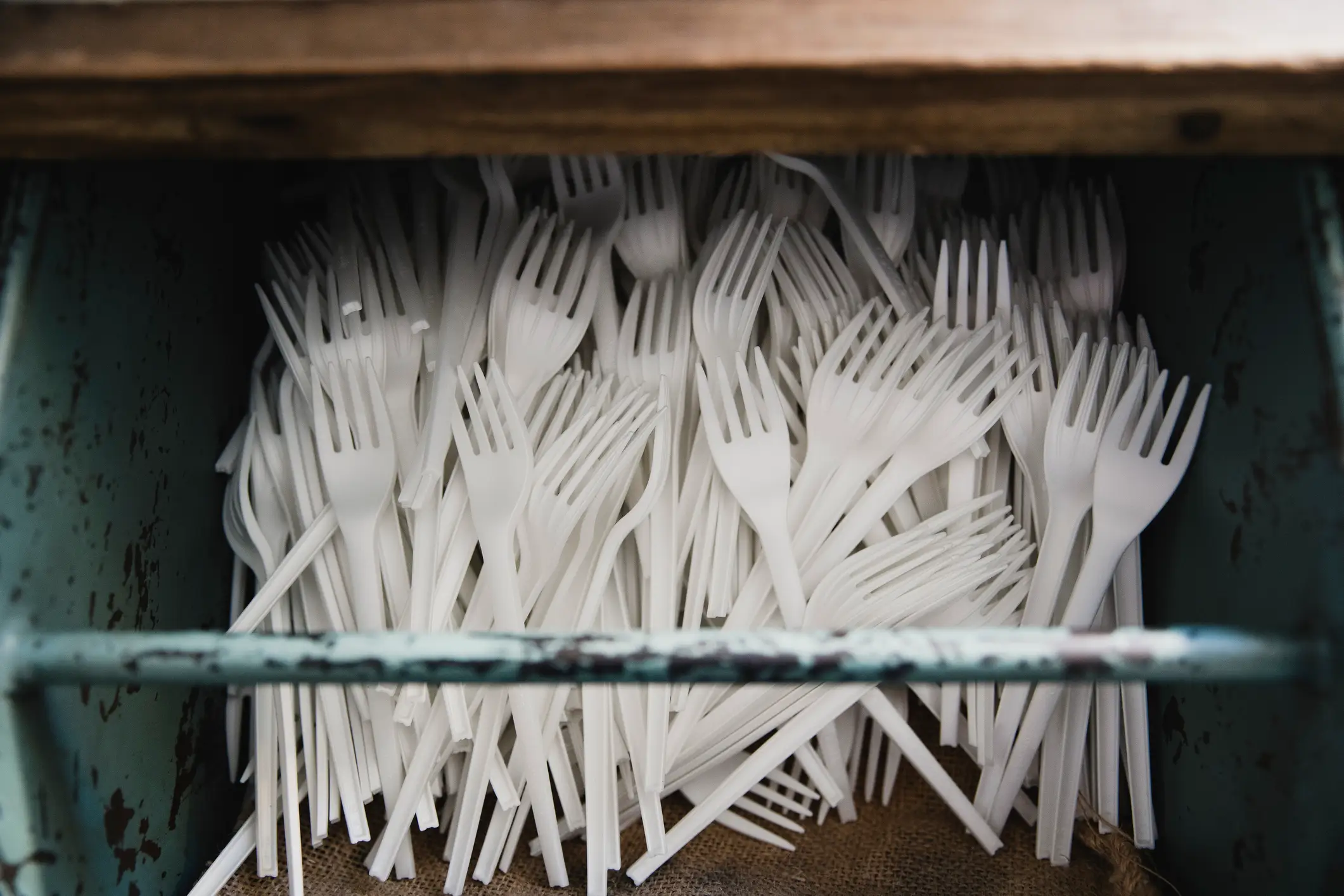 White disposable plastic forks are piled inside a rustic teal metal container beneath a wooden edge.