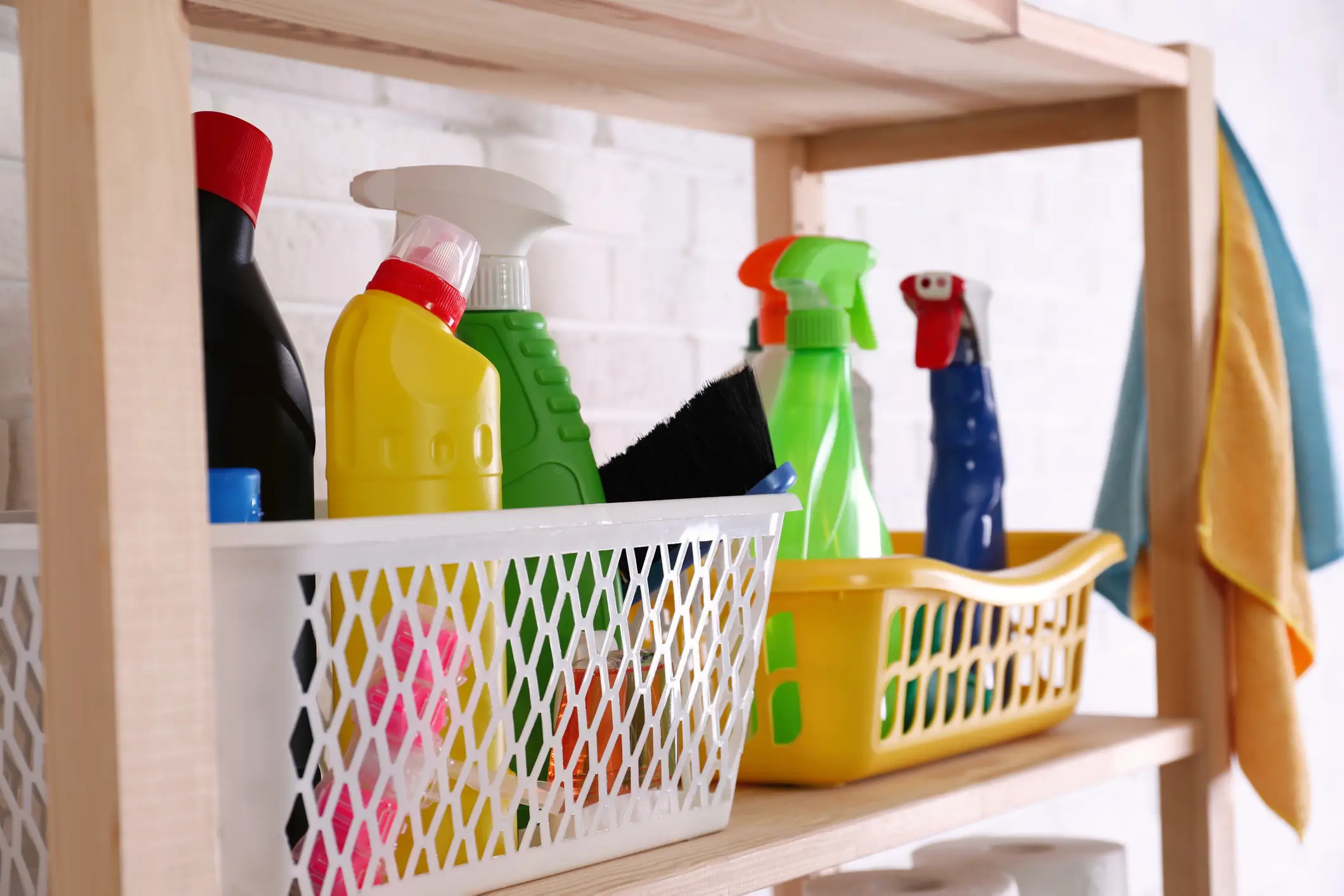 Cleaning sprays, brushes, and cloths sit in plastic baskets on a wooden shelf against a white brick wall.