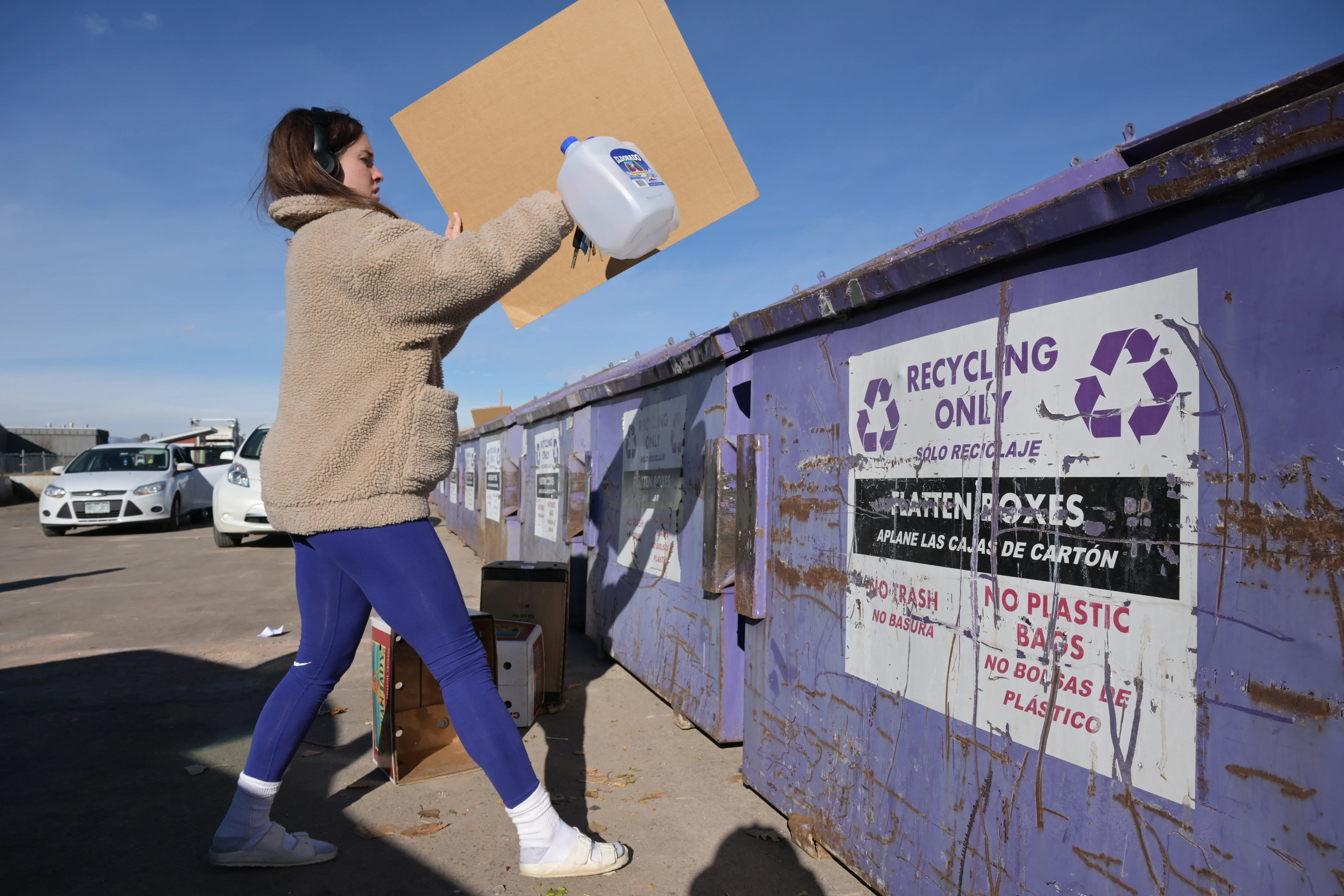 Person places a plastic jug and cardboard into a purple recycling dumpster at an outdoor collection site.