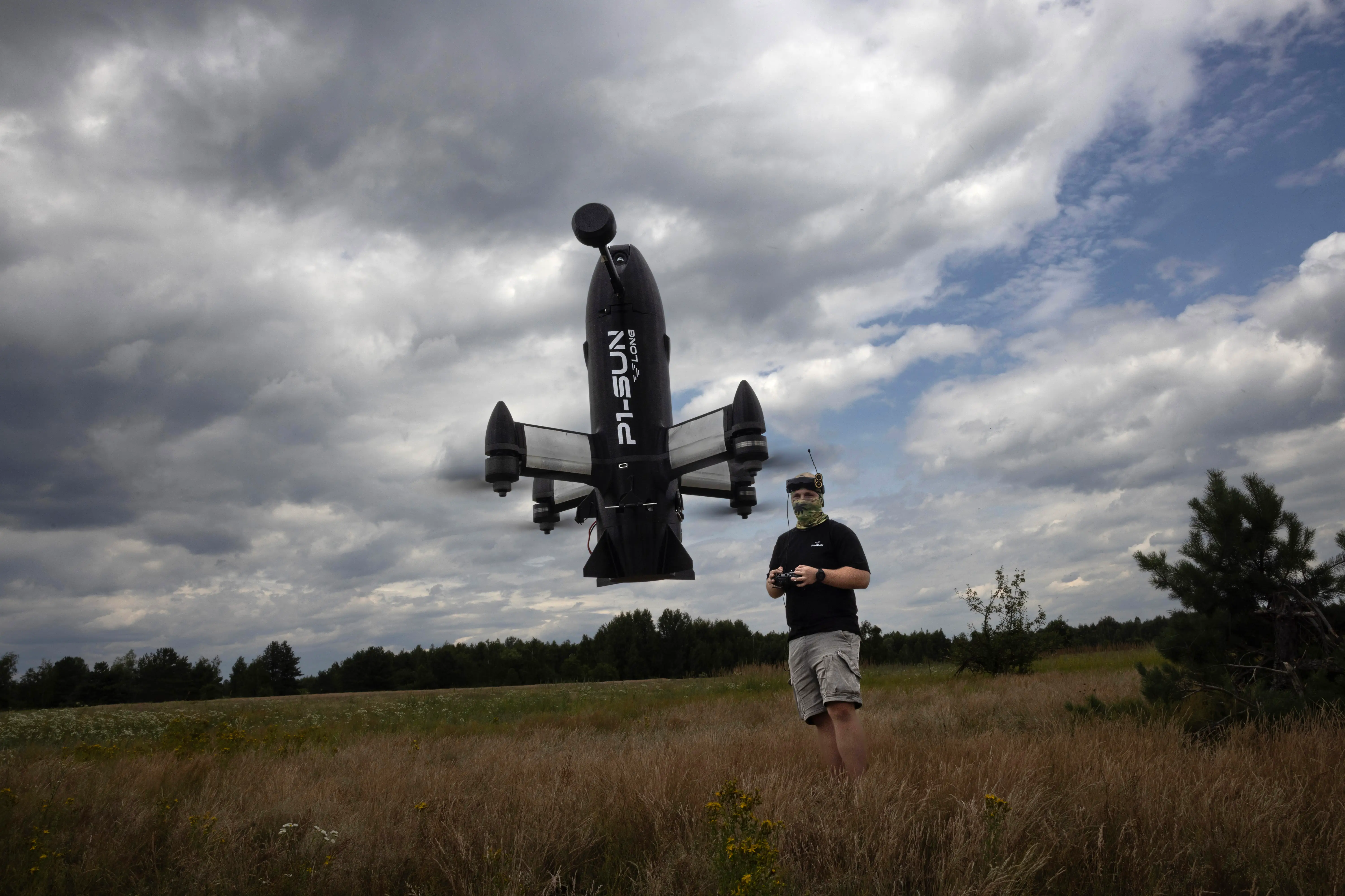 A black Pivotal VTOL aircraft hovers over a grassy field as a controller operator stands under cloudy skies.