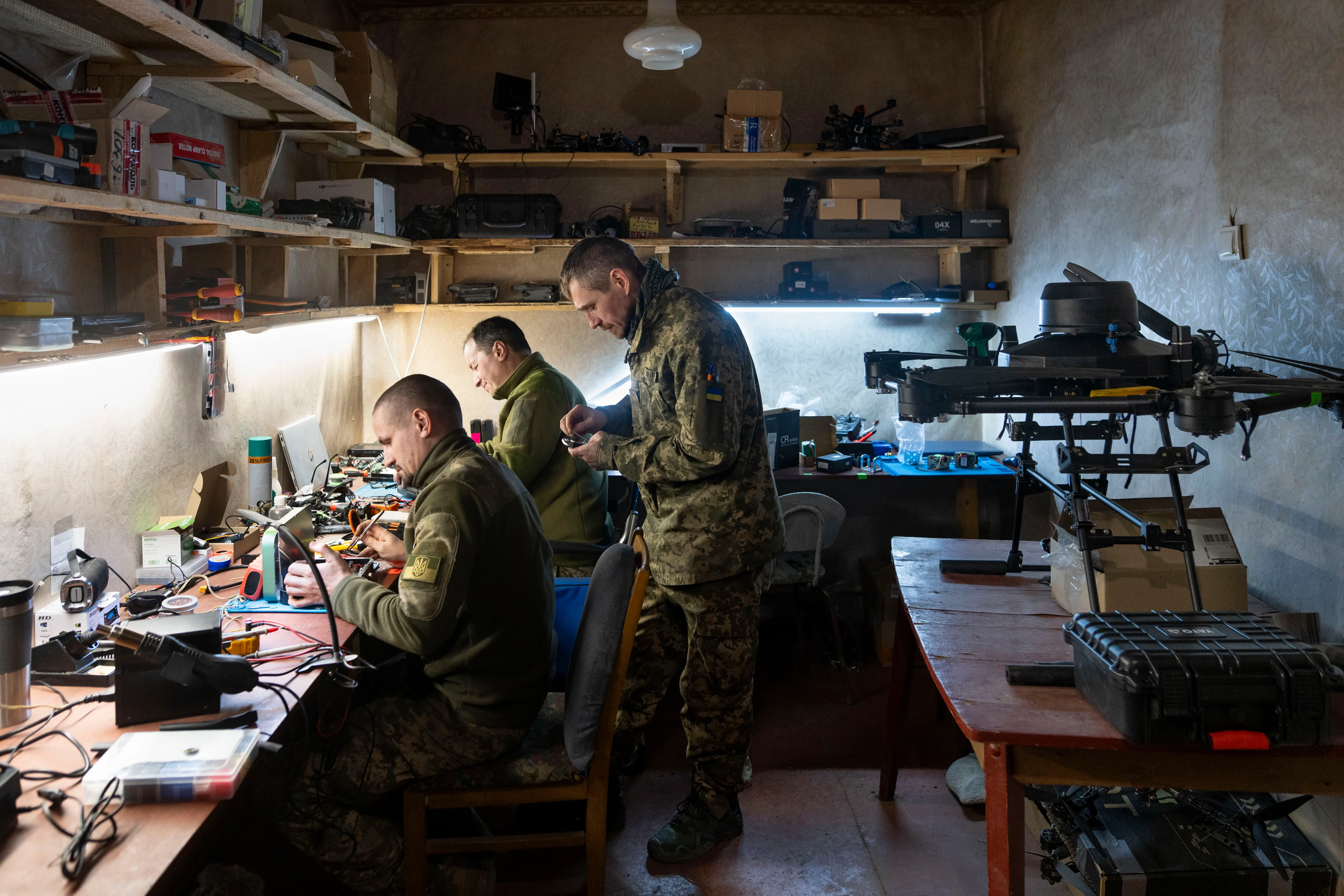 Uniformed personnel work on electronics beside a large drone in a cluttered workshop with shelves and tools.