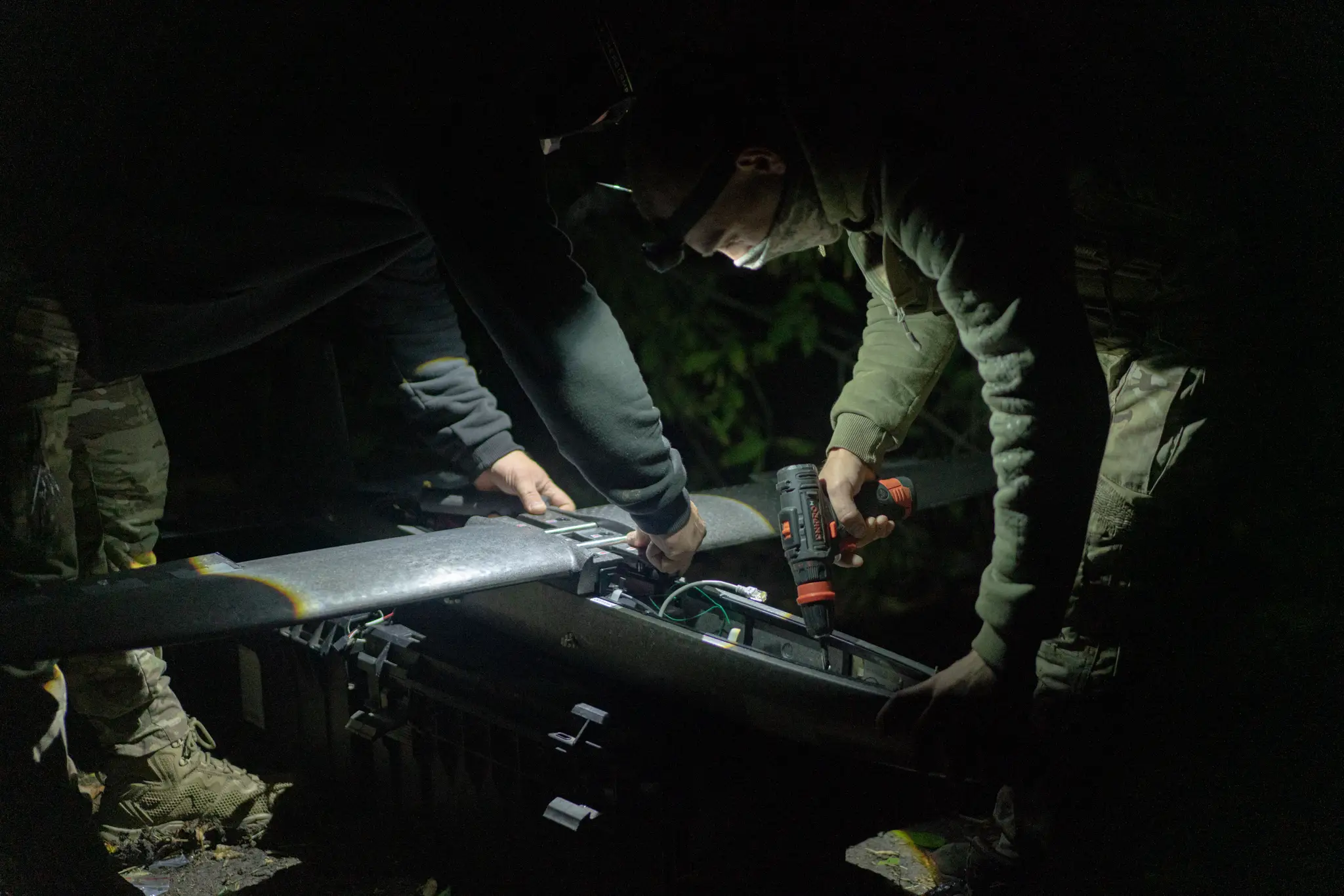 People in camouflage clothing use a drill and hand tools on a large black aircraft component at night.