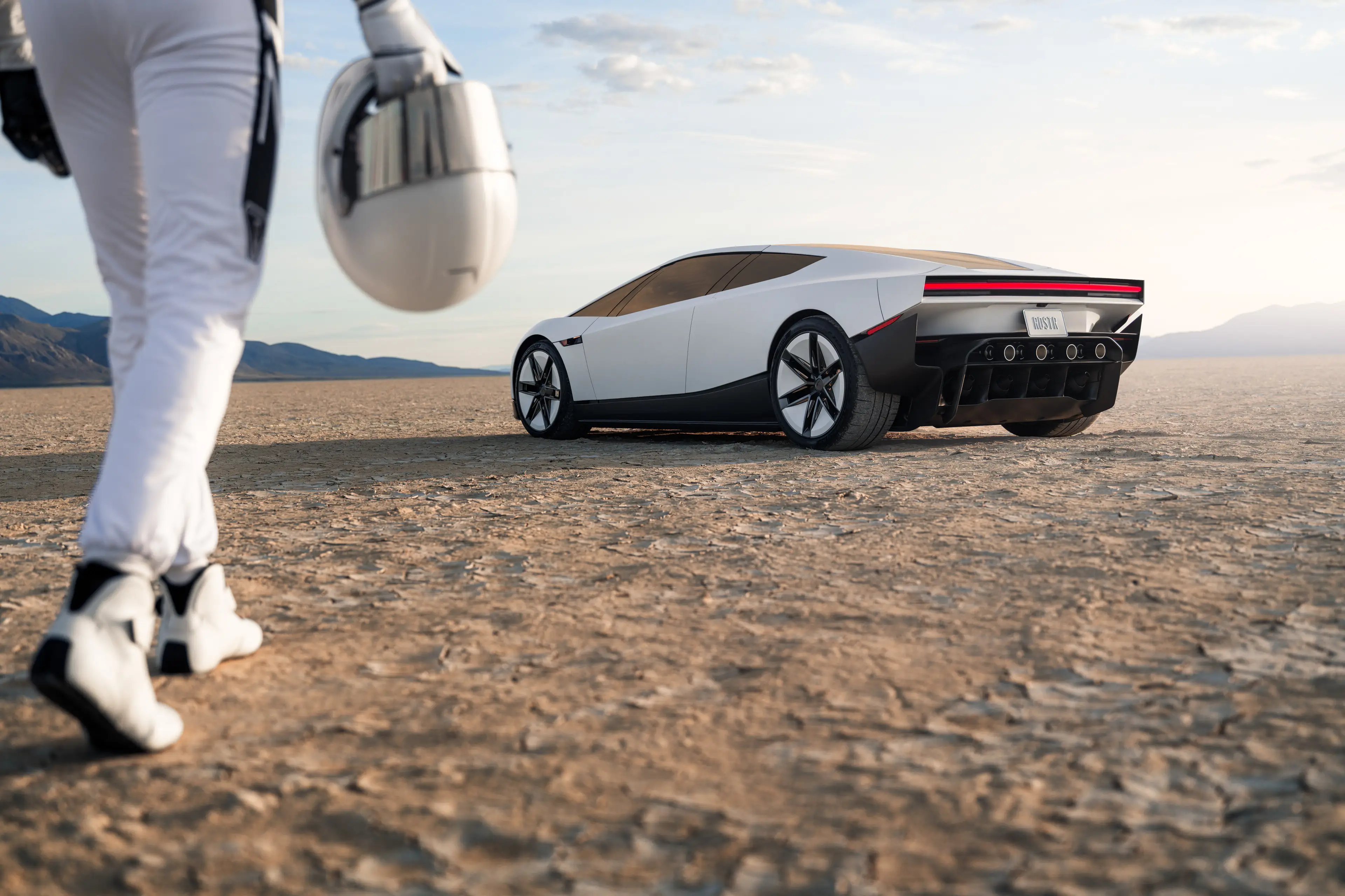 A person in a white outfit holding a helmet is walking toward a Tesla Roadster.