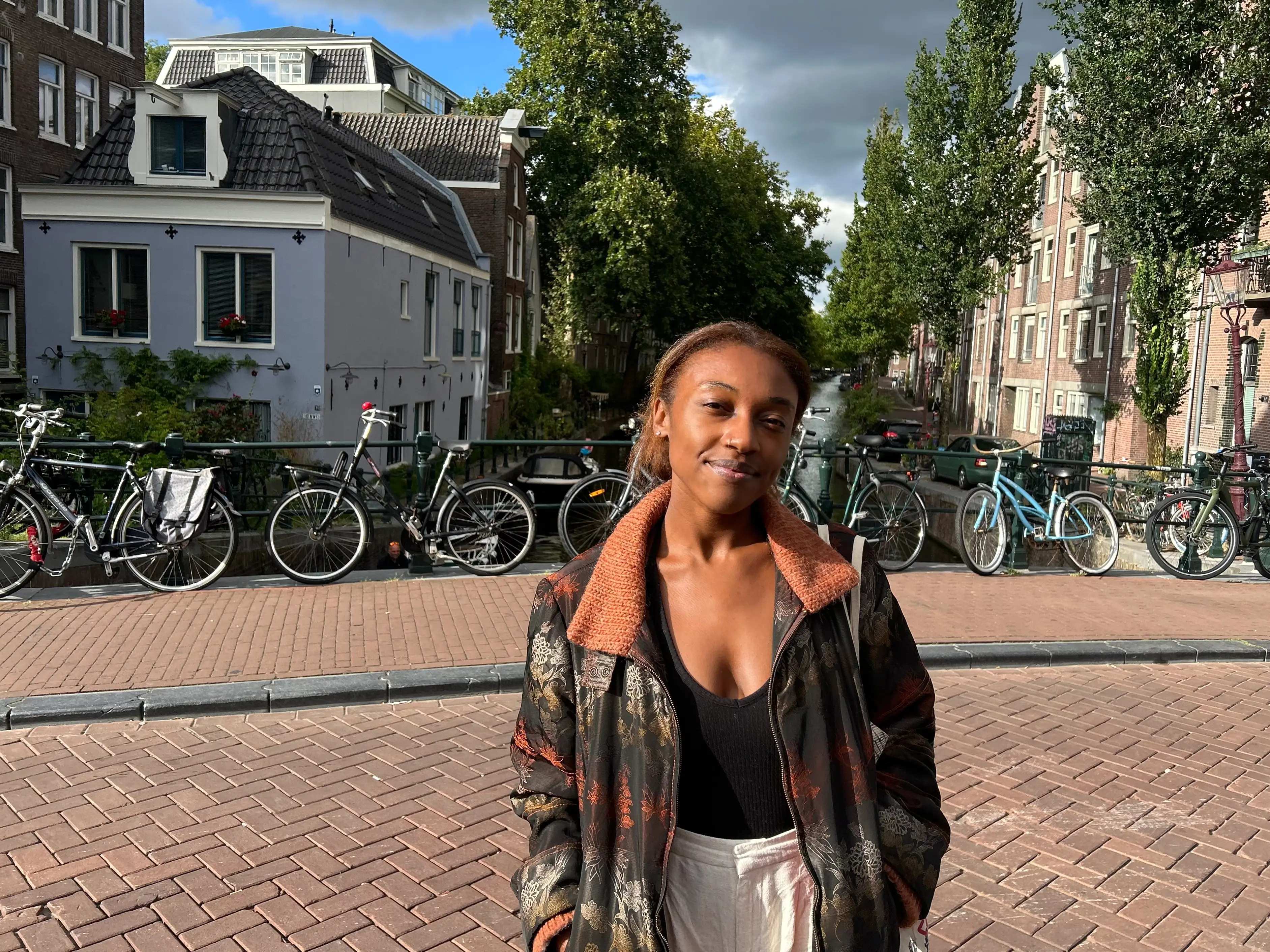 The writer stands on a brick canal bridge lined with bicycles, trees, and urban residential buildings.