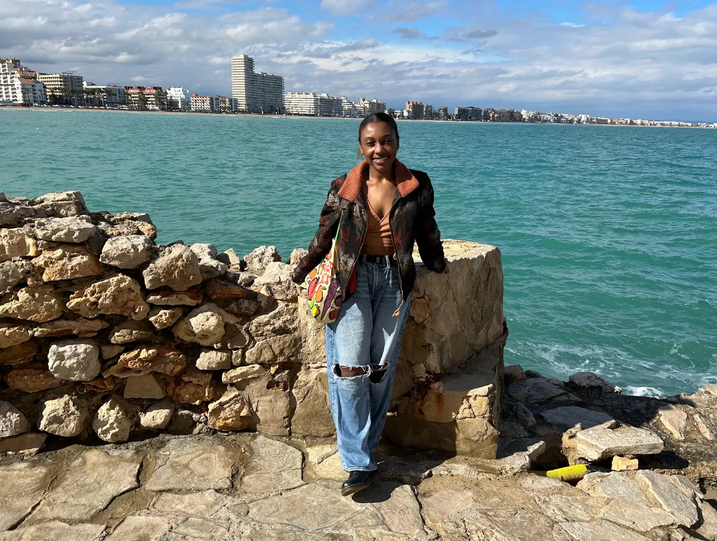 The writer stands by a stone seawall overlooking turquoise water and a coastal city skyline.