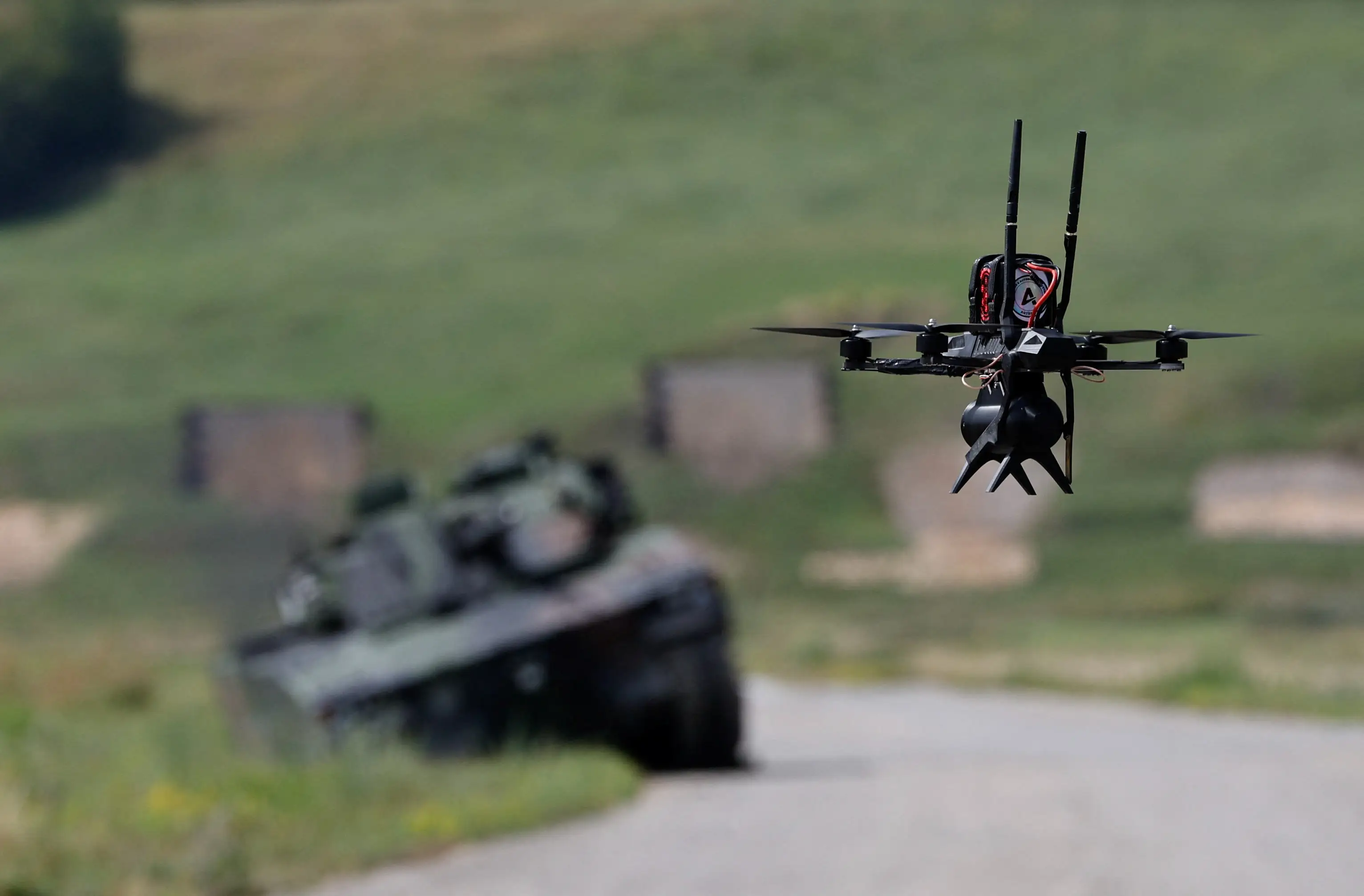 Quadcopter drone flies above a road with a blurred armored vehicle in a grassy training area.