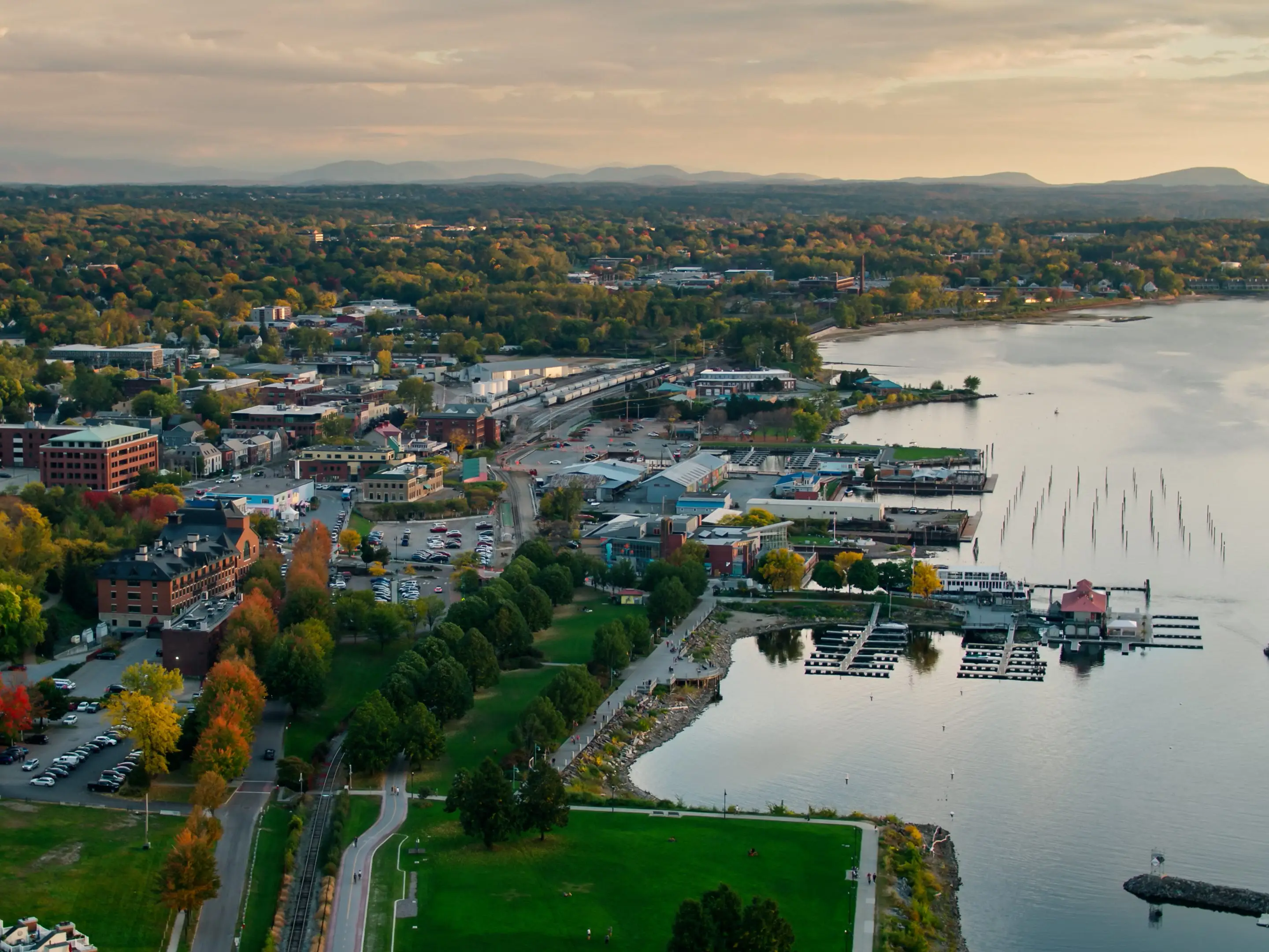 Drone shot of the shore of Lake Champlain in Burlington, Vermont at sunset, including the ferry terminal, commercial docks, and downtown streets shaded by trees that are beginning to to turn red, yellow and orange. The Green Mountains can be seen in the distance. Authorization was obtained from the FAA for this operation in restricted airspace.