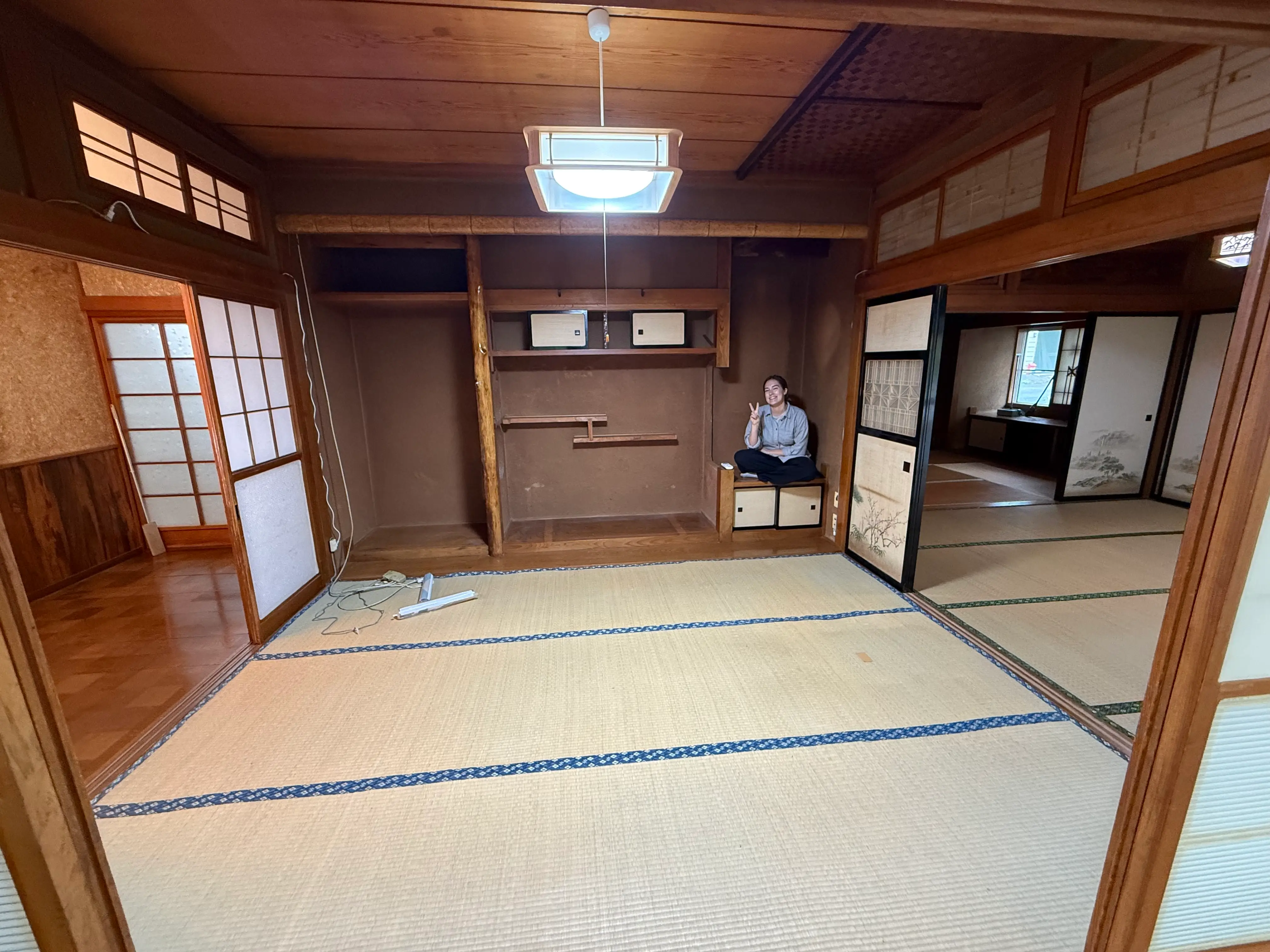 A person sits in a traditional tatami room with wooden sliding doors, built-in shelves, and a ceiling light.