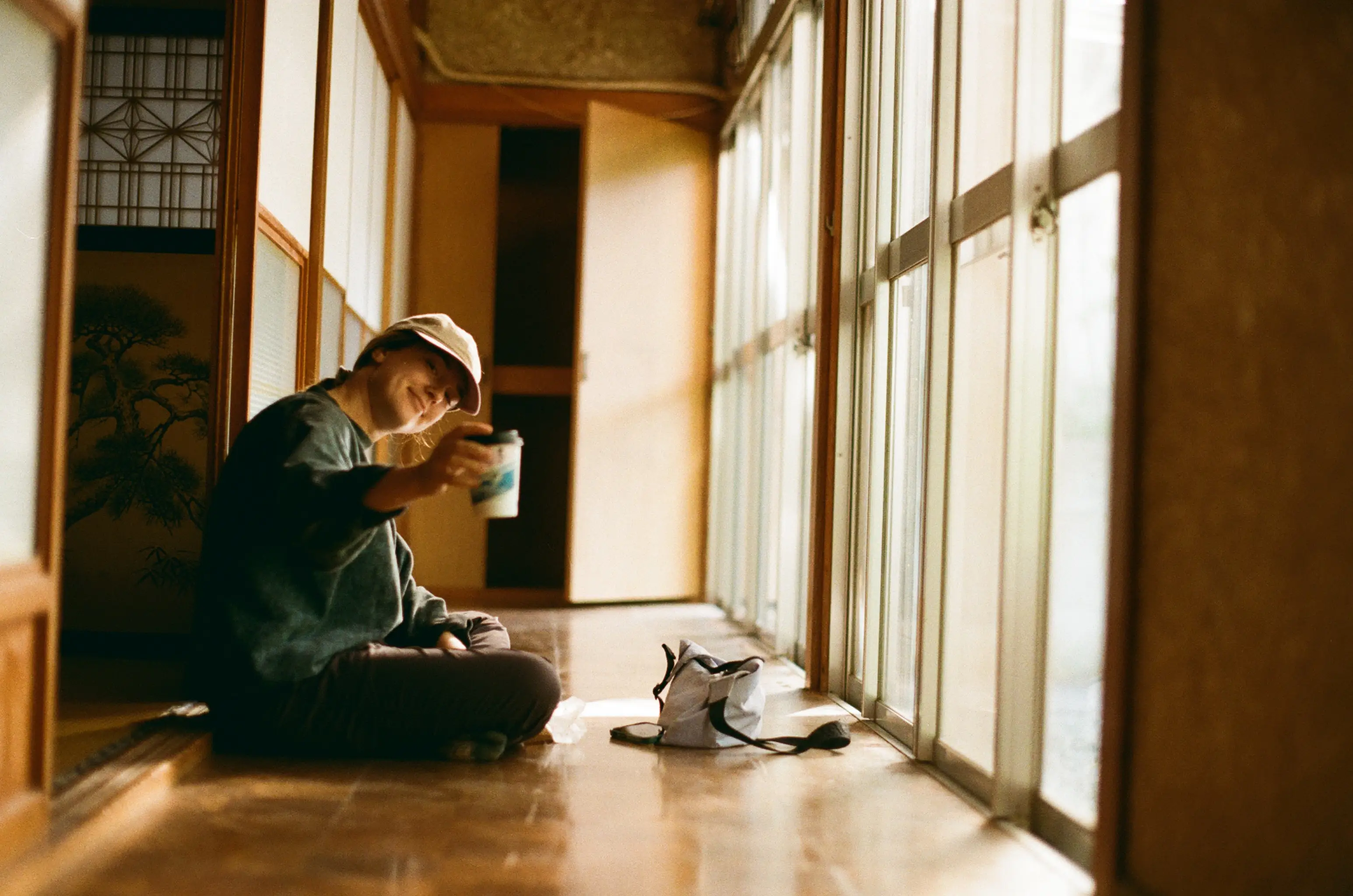 Person sits on a polished wooden corridor floor holding a cup beside sunlit sliding windows.