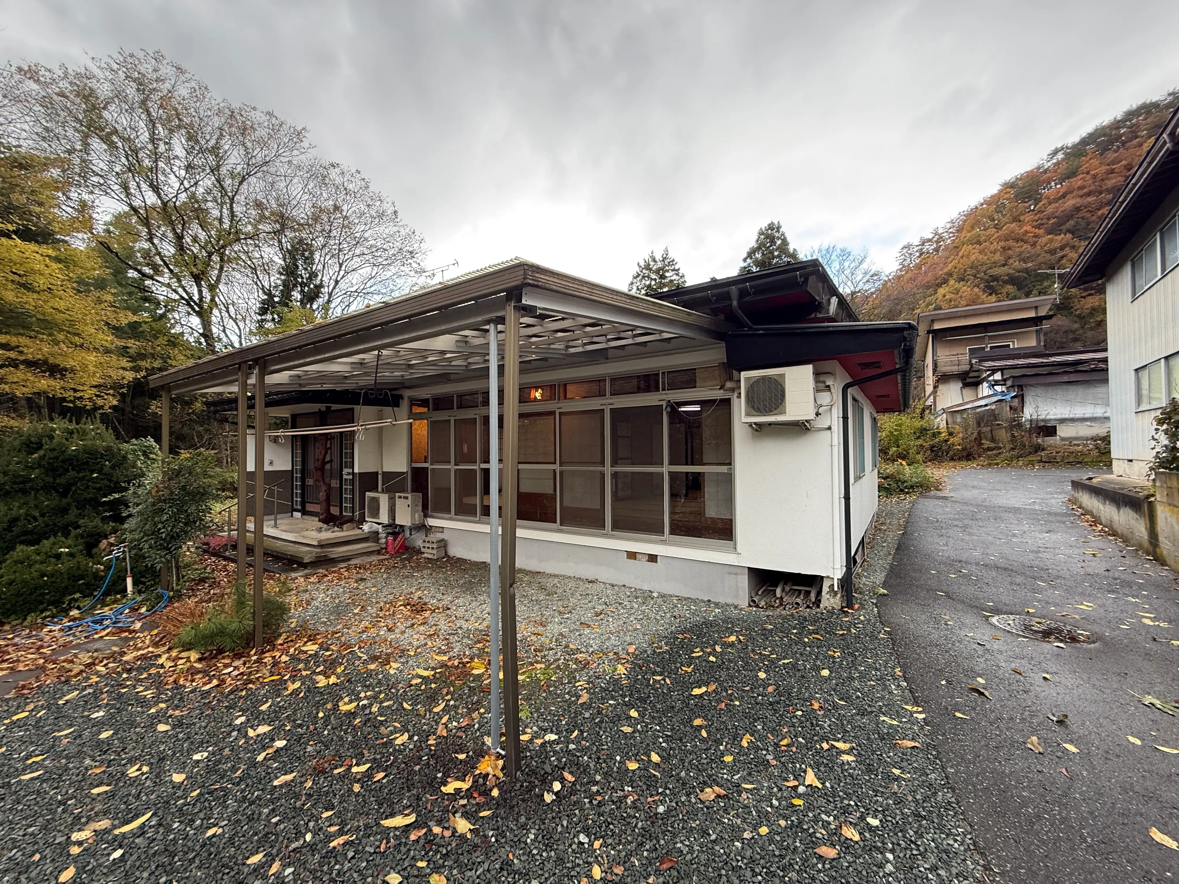A small house with a covered porch stands beside a driveway under cloudy skies and autumn trees.