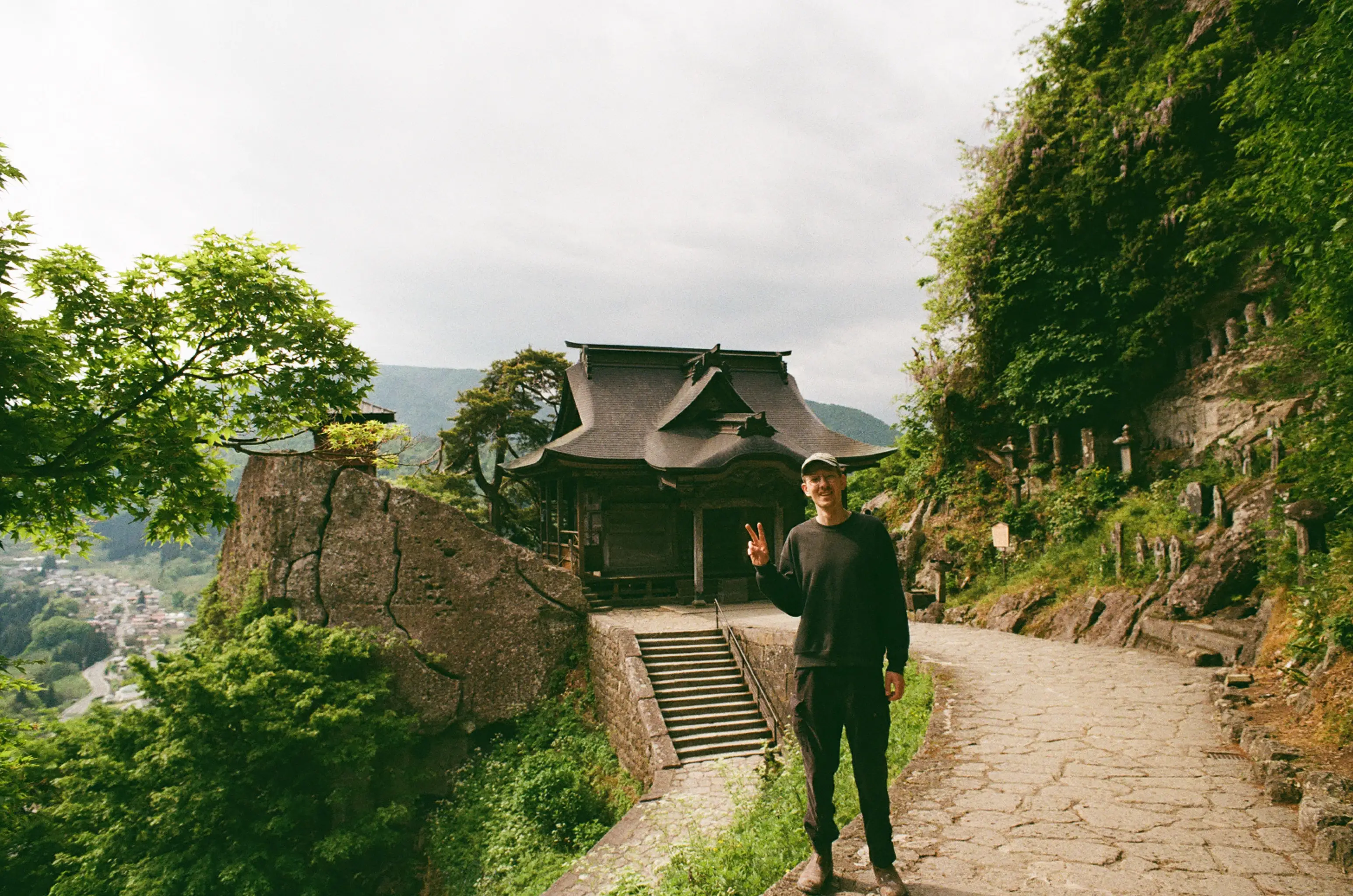 Person flashes a peace sign on a stone path beside a traditional temple building in a lush mountainside setting.