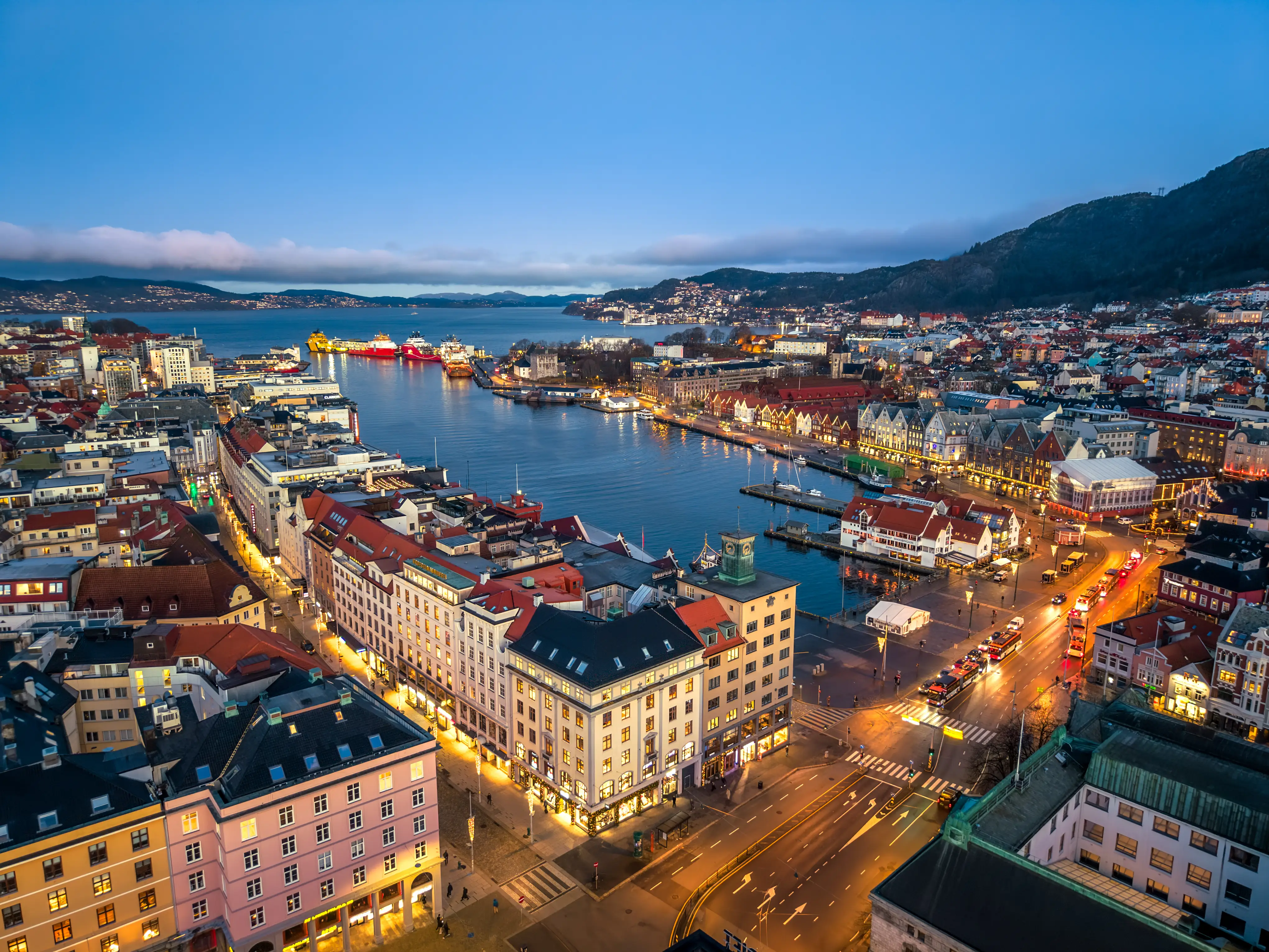 Twilight aerial view of Bergen harbor with illuminated waterfront buildings, boats, roads, and mountains surrounding the bay.