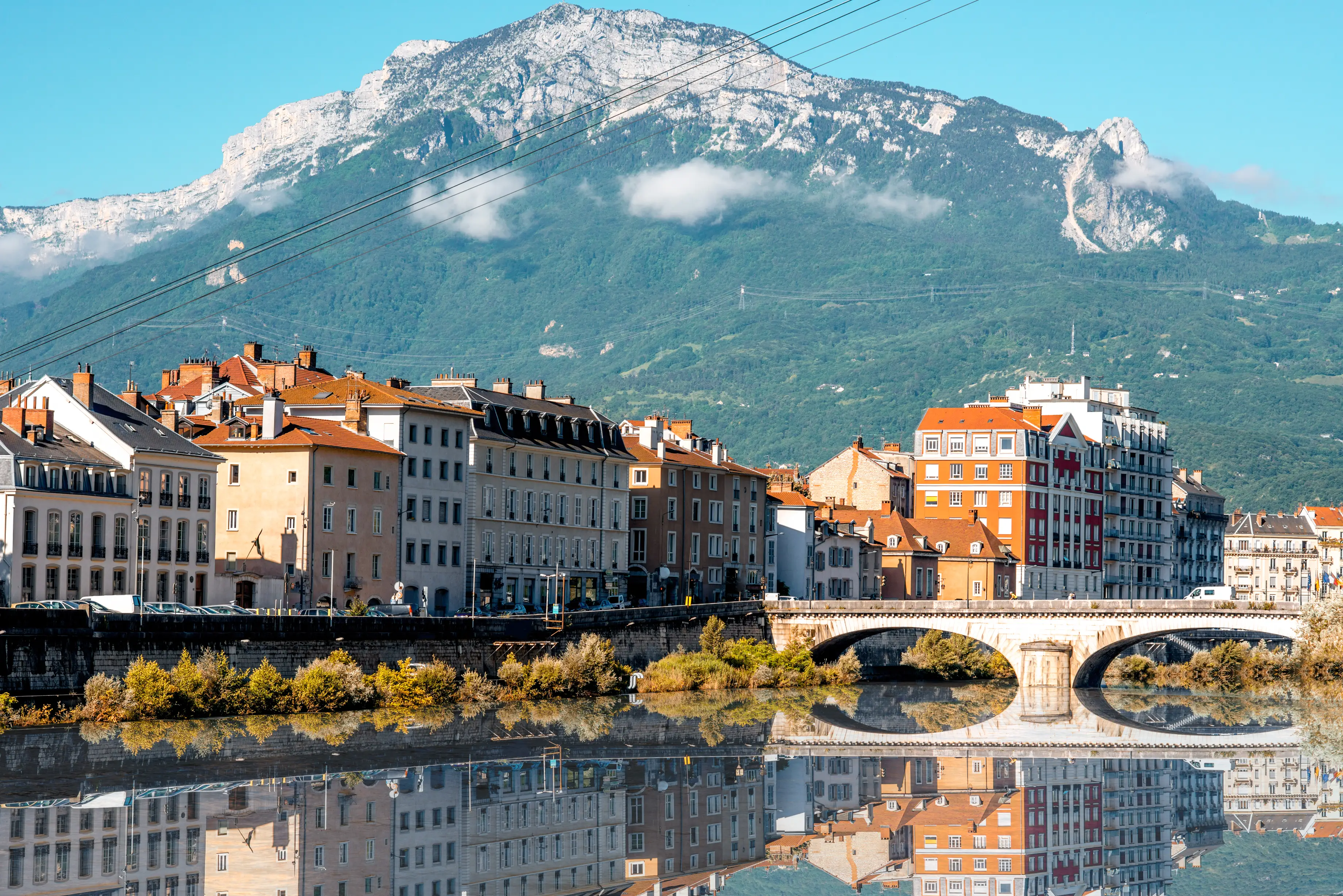 Riverside buildings and an arched stone bridge reflect in calm water beneath a snow-capped mountain.