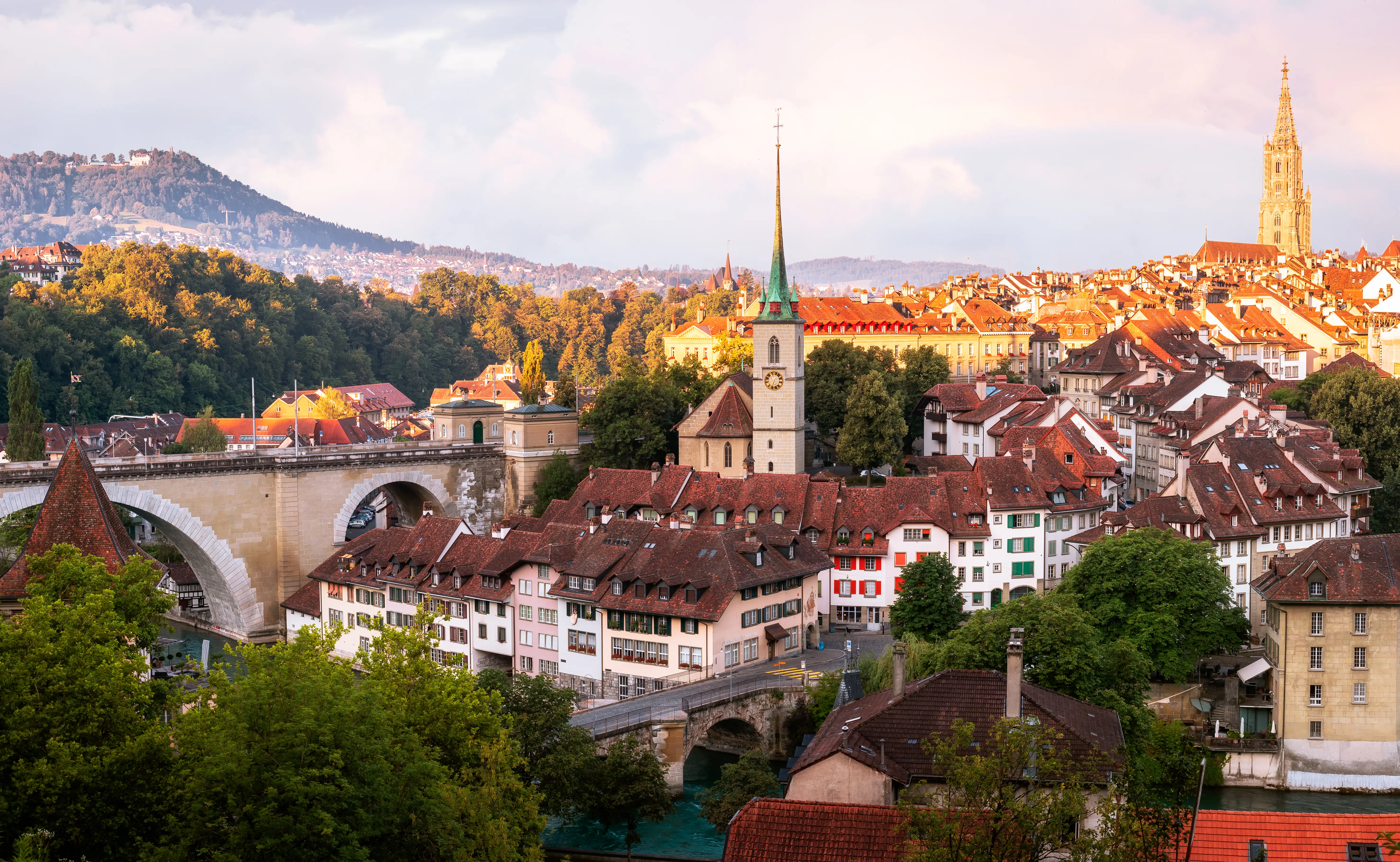 Bern old town rooftops, church spires, stone bridge, and hillside are lit by warm evening sunlight.