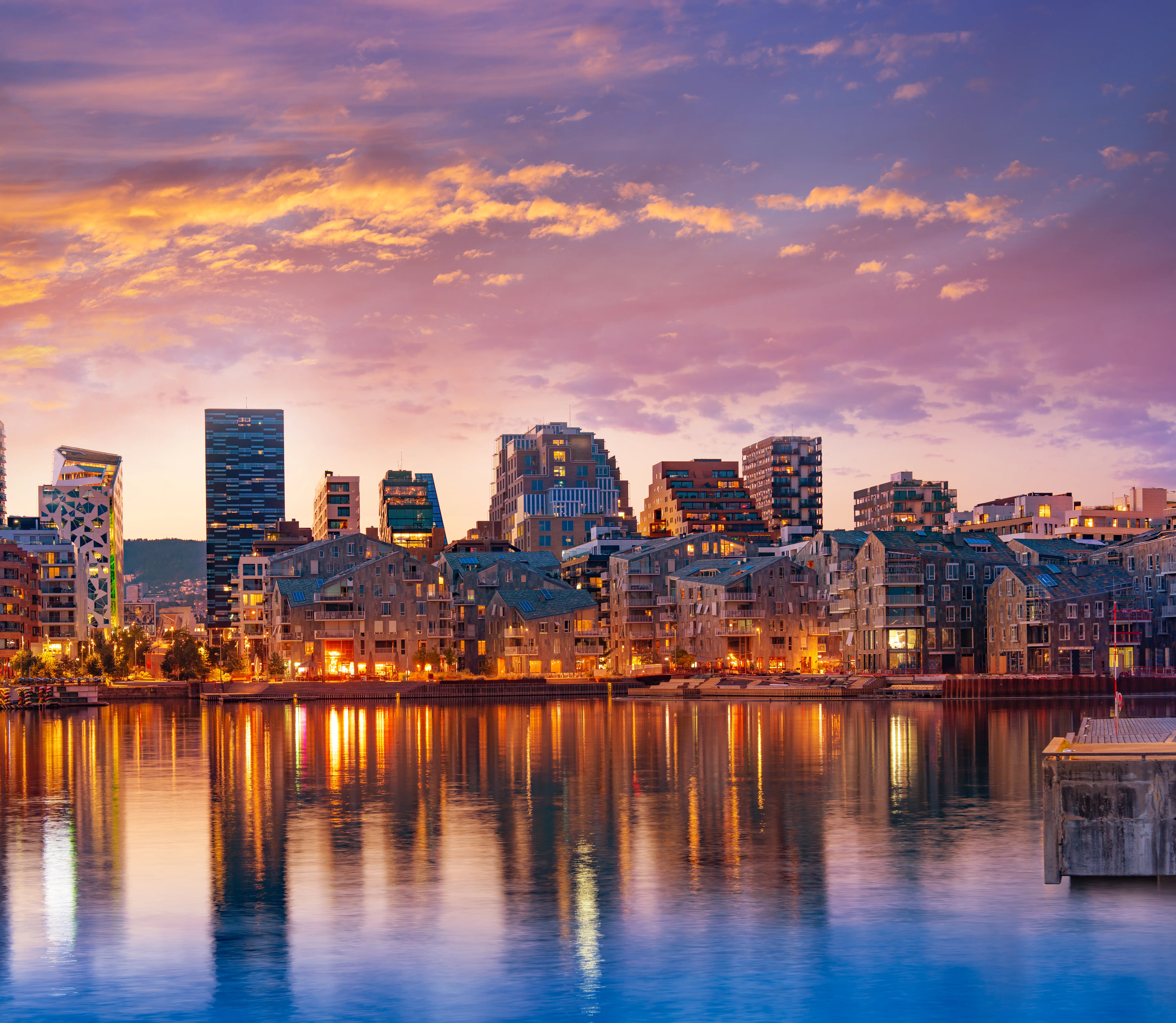 Modern waterfront skyline glows at sunset, with city lights reflecting across calm harbor water.