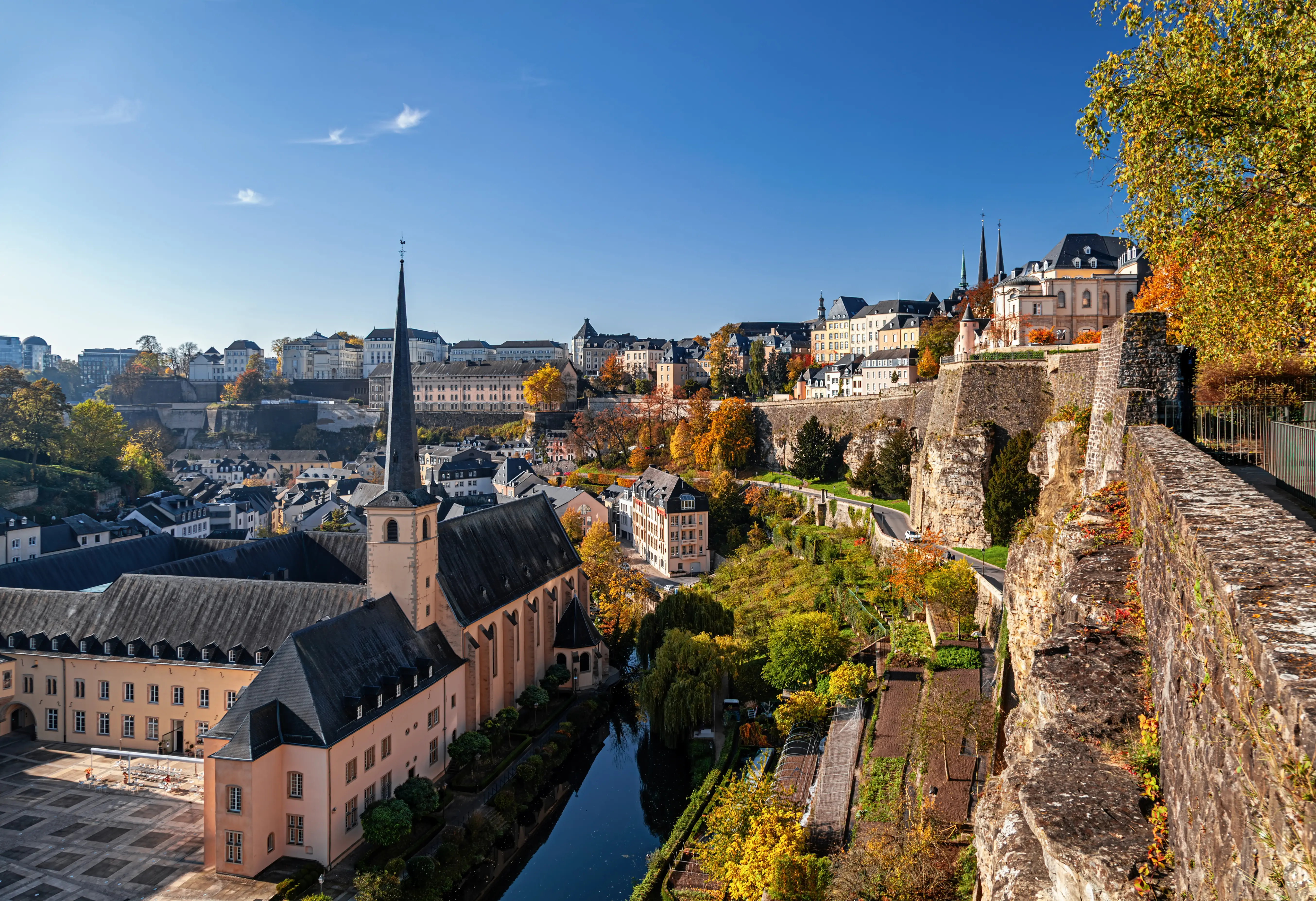 Luxembourg City old town overlooks a church, canal, stone ramparts, and autumn trees under a clear blue sky.