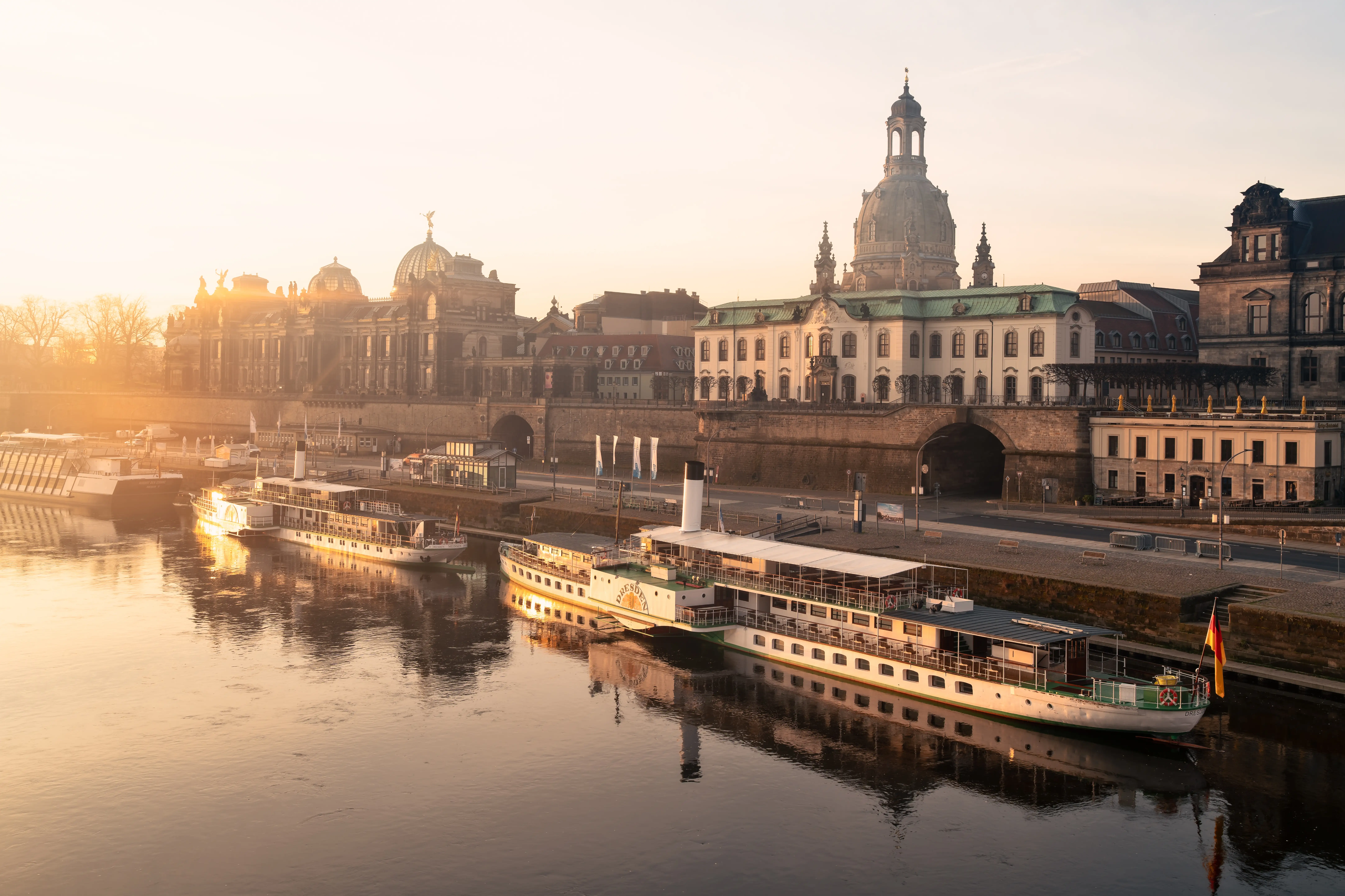 River cruise boats sit on the Elbe beside Dresden’s historic waterfront and domed skyline at sunset.