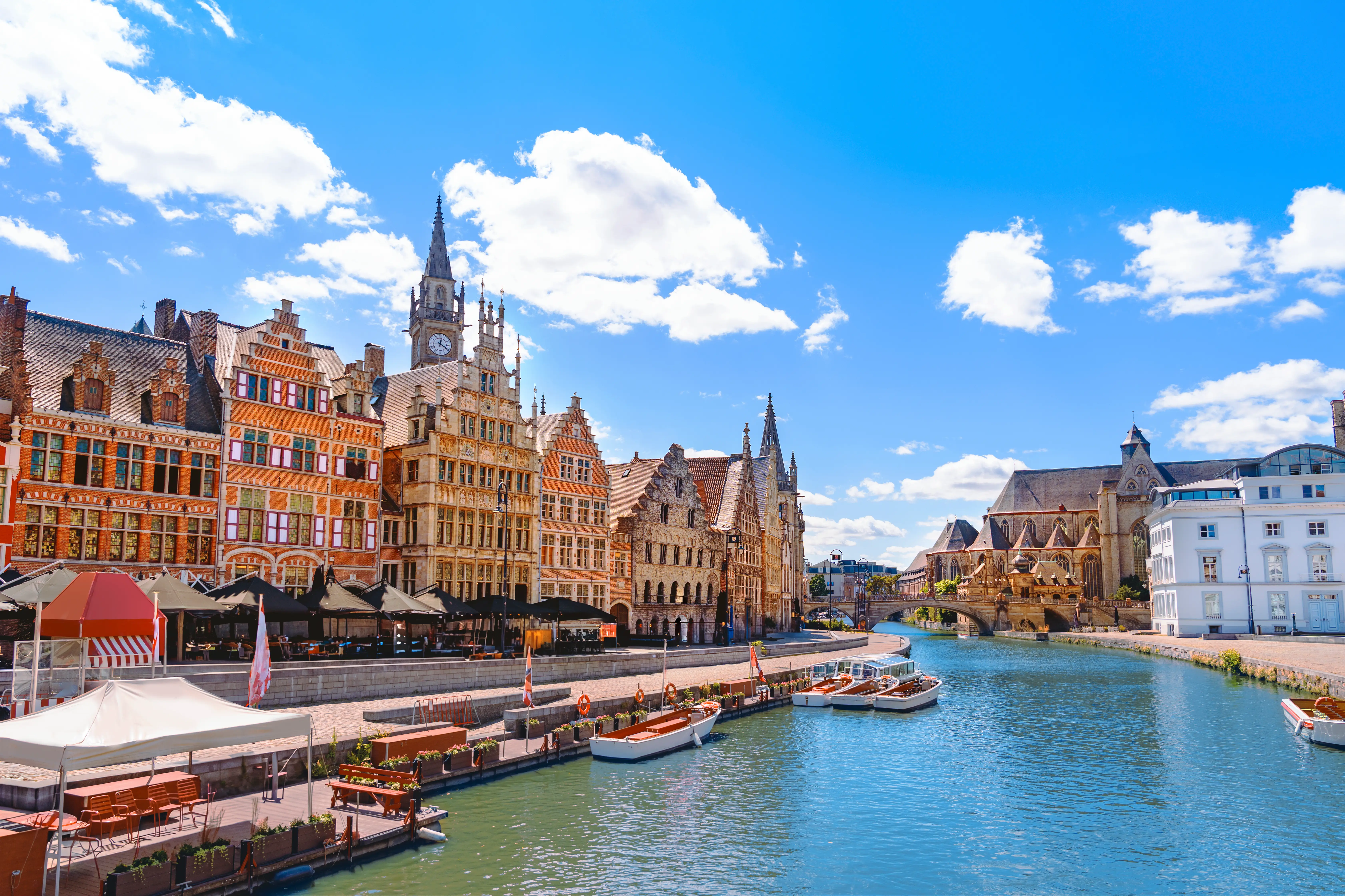 Historic riverside buildings line a canal with boats under a bright blue sky in Ghent, Belgium.