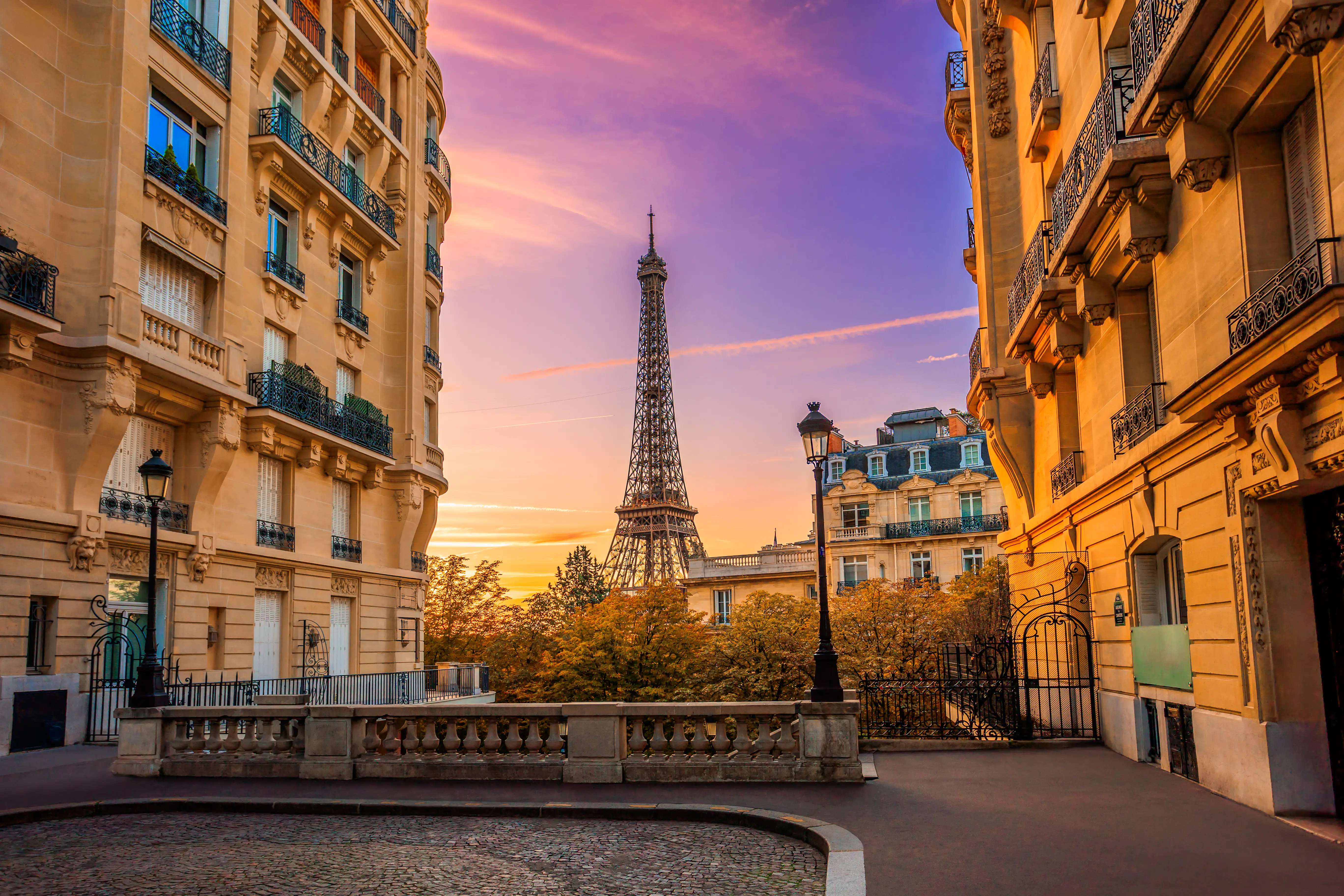 Eiffel Tower rises between Paris apartment buildings under a colorful sunset sky.