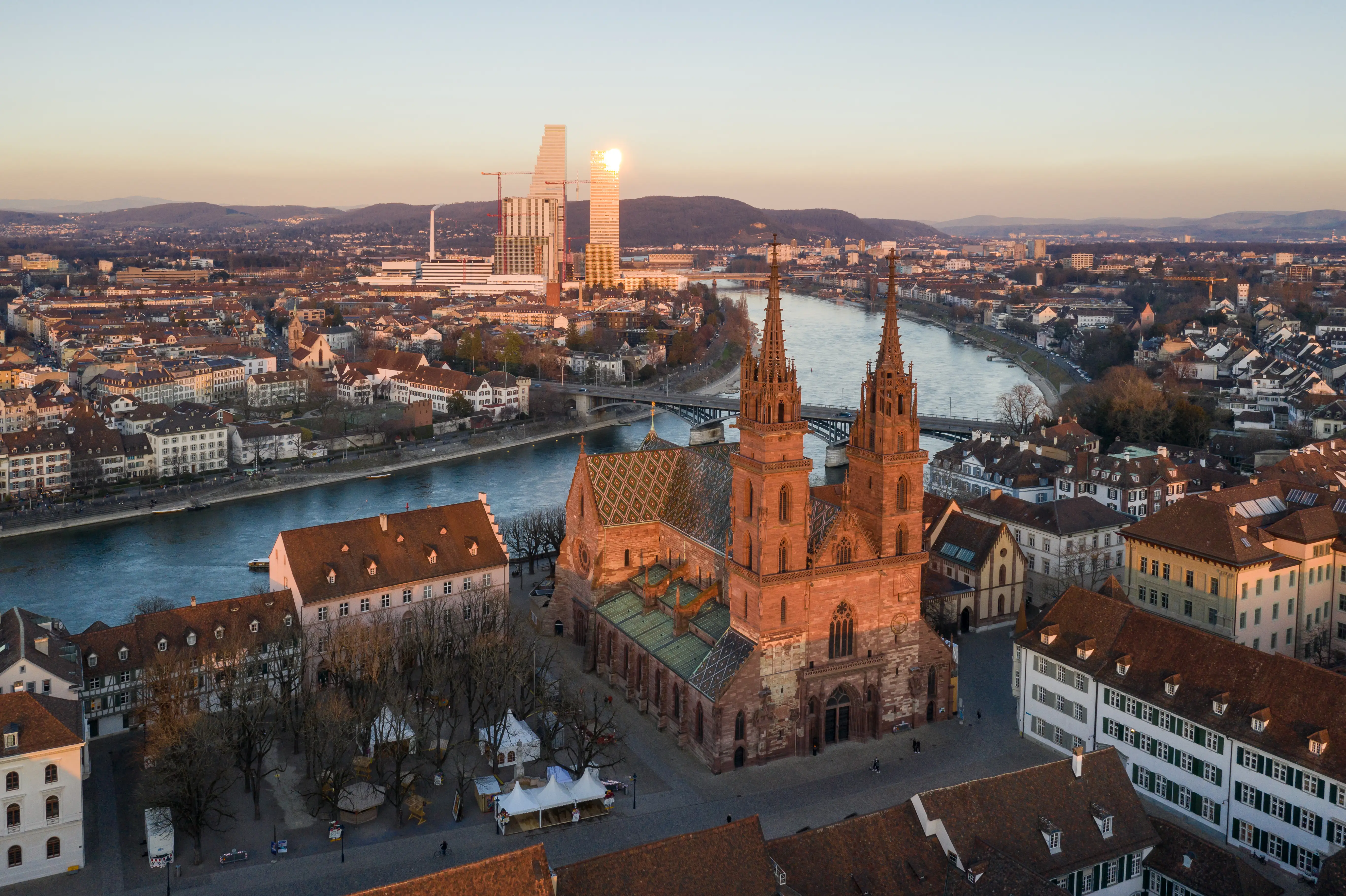 Aerial view of Basel Minster beside the Rhine River at sunset, with city rooftops and towers in the background.