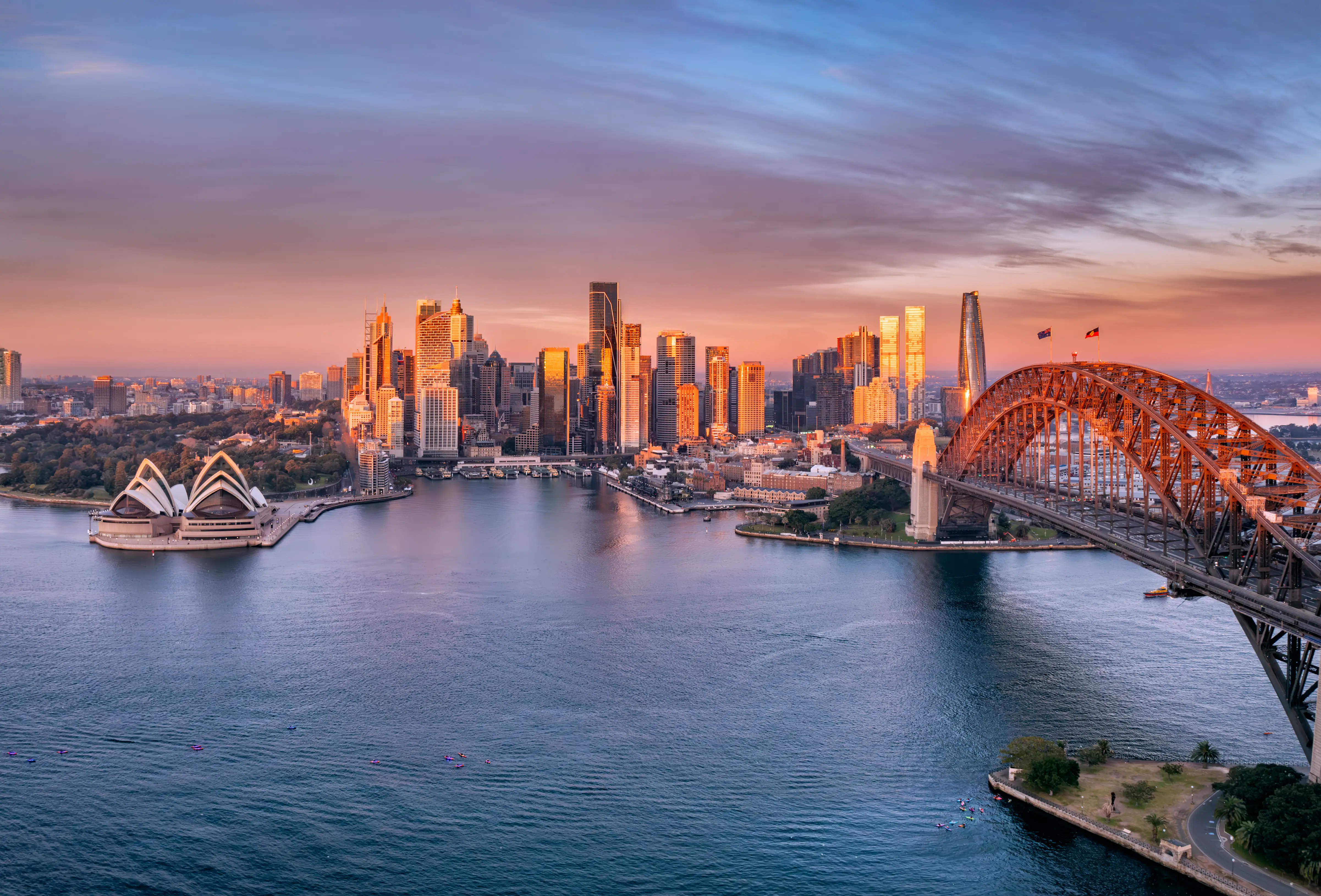 Sydney Harbour at sunset with the Opera House, Harbour Bridge, and illuminated downtown skyline.