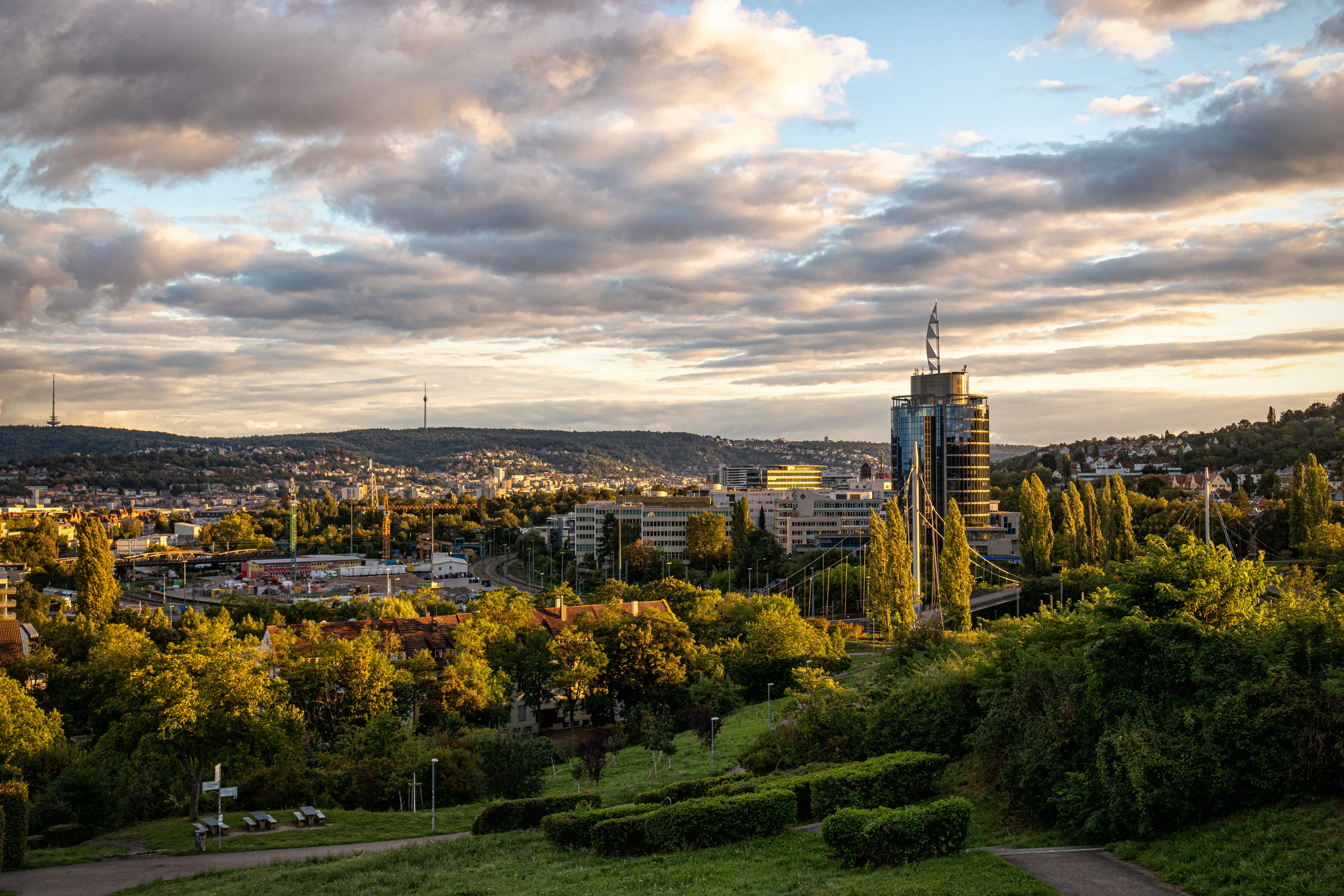 Sunset city skyline with modern high-rise buildings, wooded hills, and a landscaped park in the foreground.