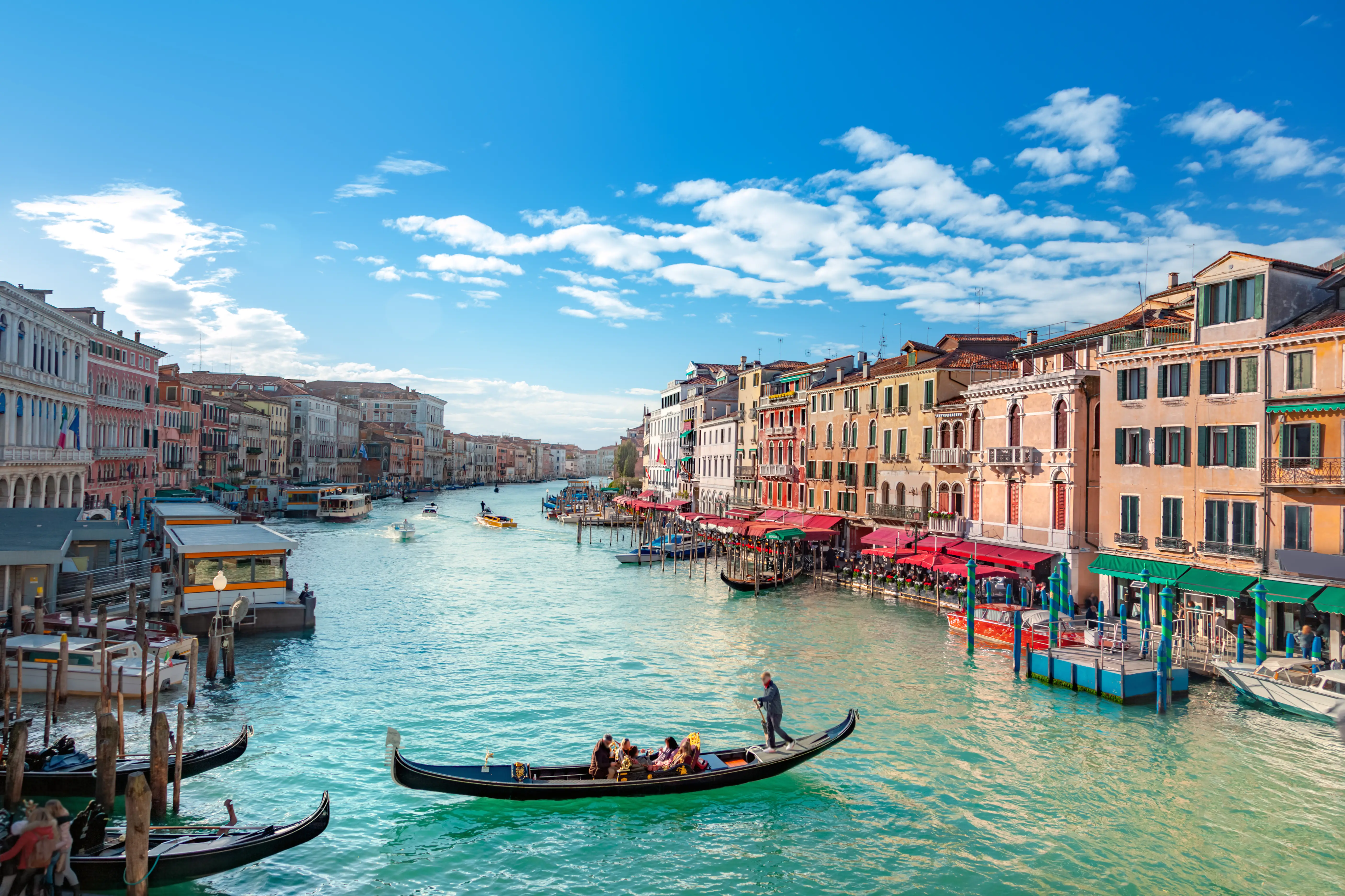 Gondolas travel along Venice's Grand Canal between colorful historic buildings under a bright blue sky.