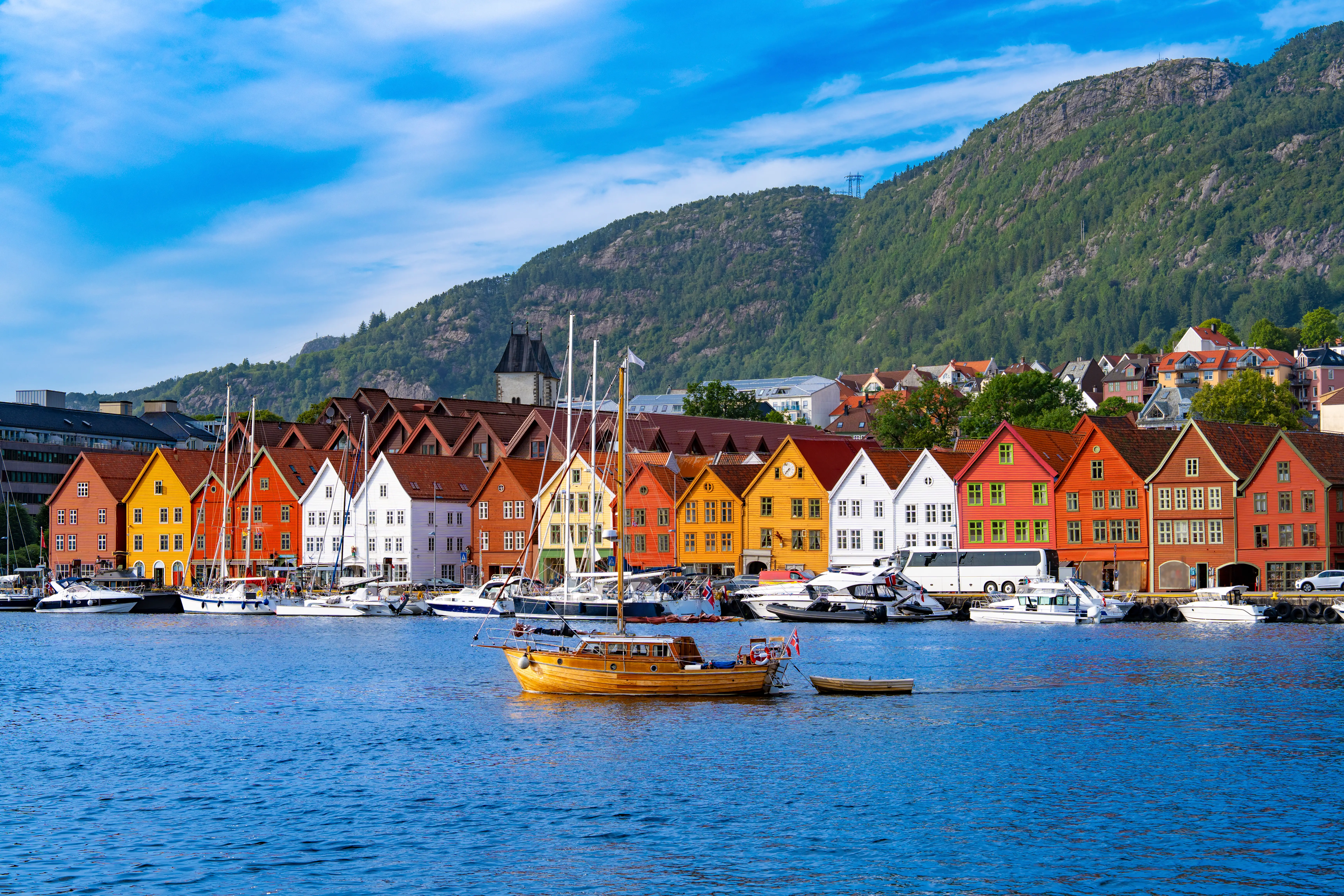 Colorful Bryggen waterfront buildings and moored boats line a harbor beneath forested mountains in Bergen, Norway.