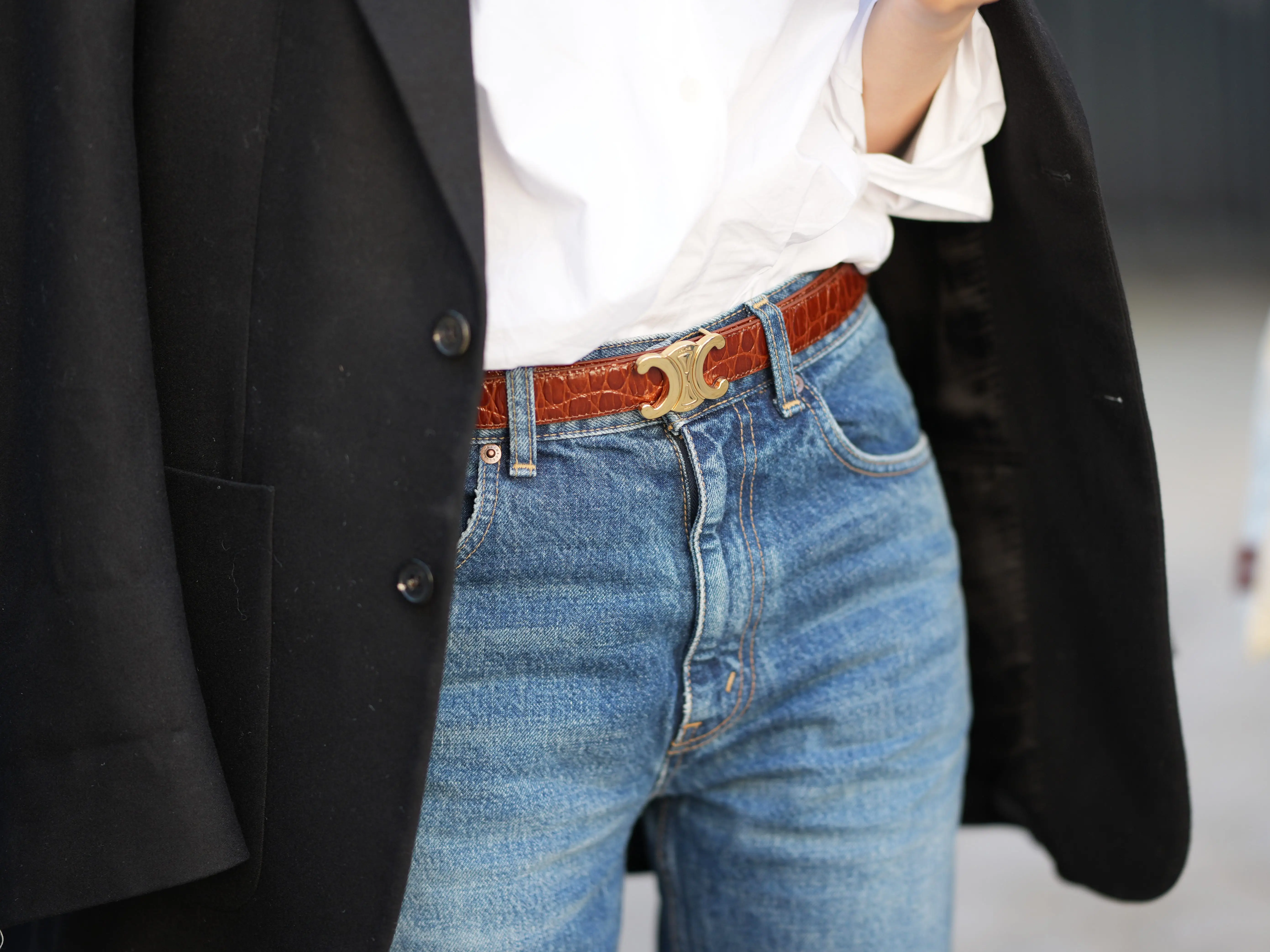 A close-up of jeans with a brown logo belt, white shirt, and black coat in a street-style outfit.
