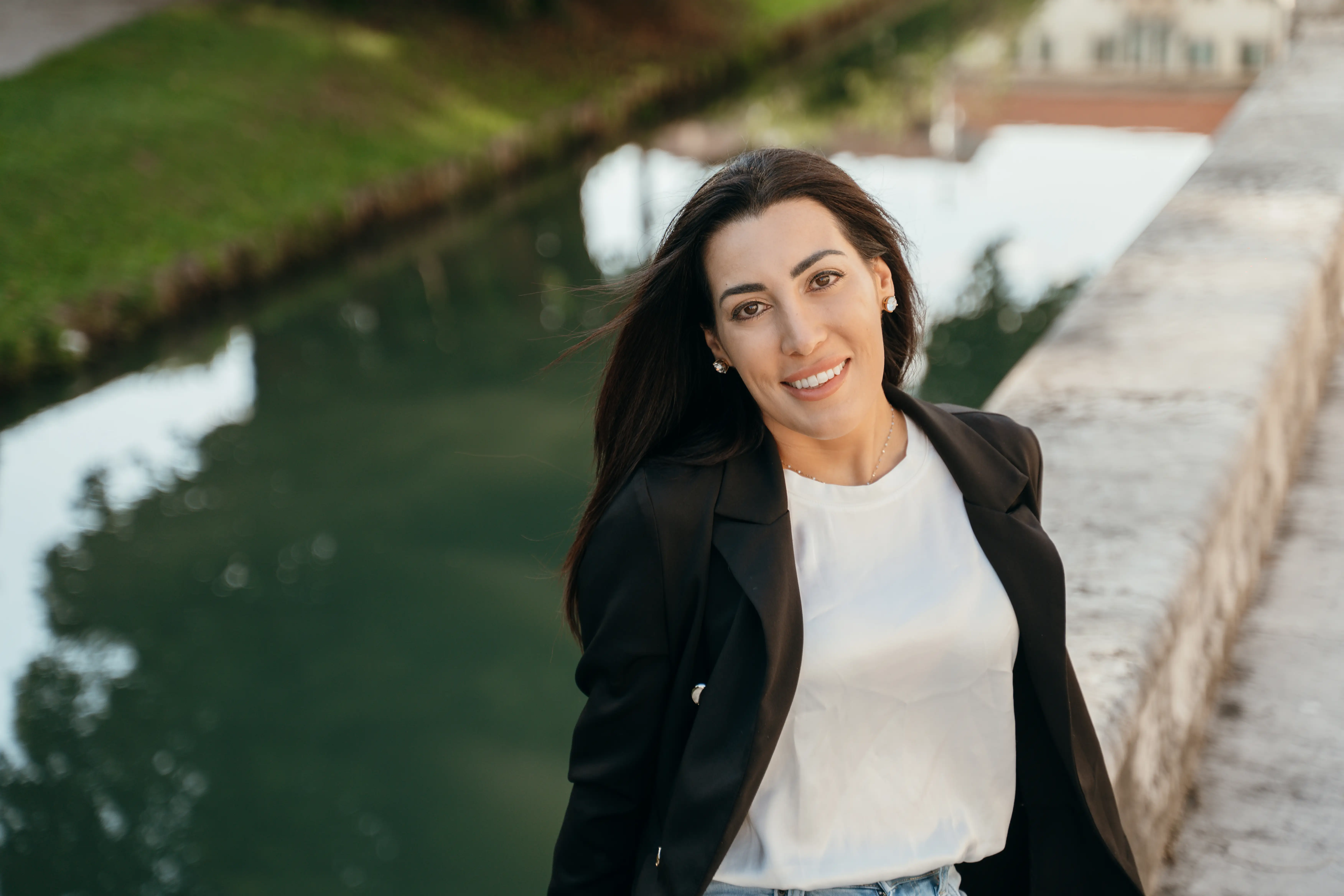 Smiling person in a black blazer stands beside a stone walkway overlooking a calm canal.
