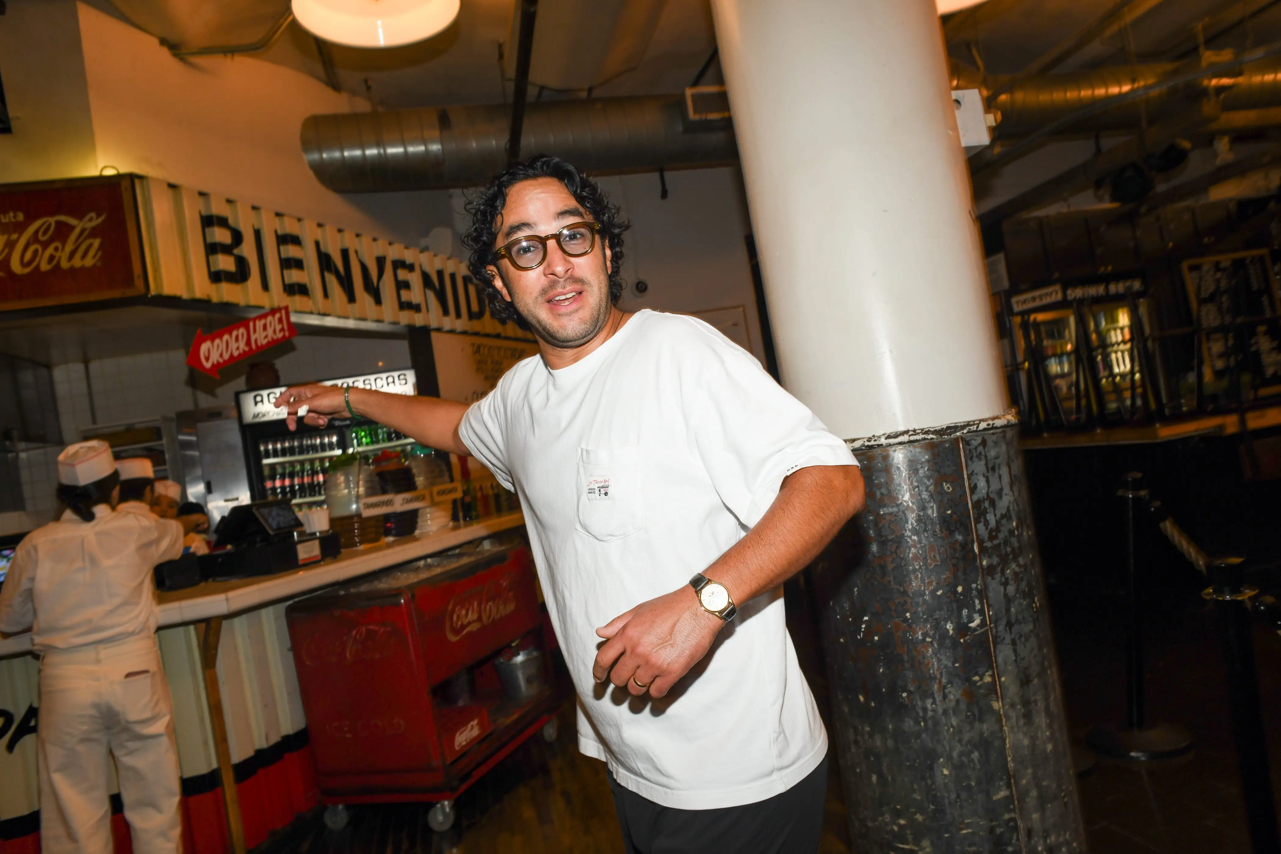 Person in glasses and a white shirt stands by a counter inside a colorful restaurant with Coca-Cola signage.