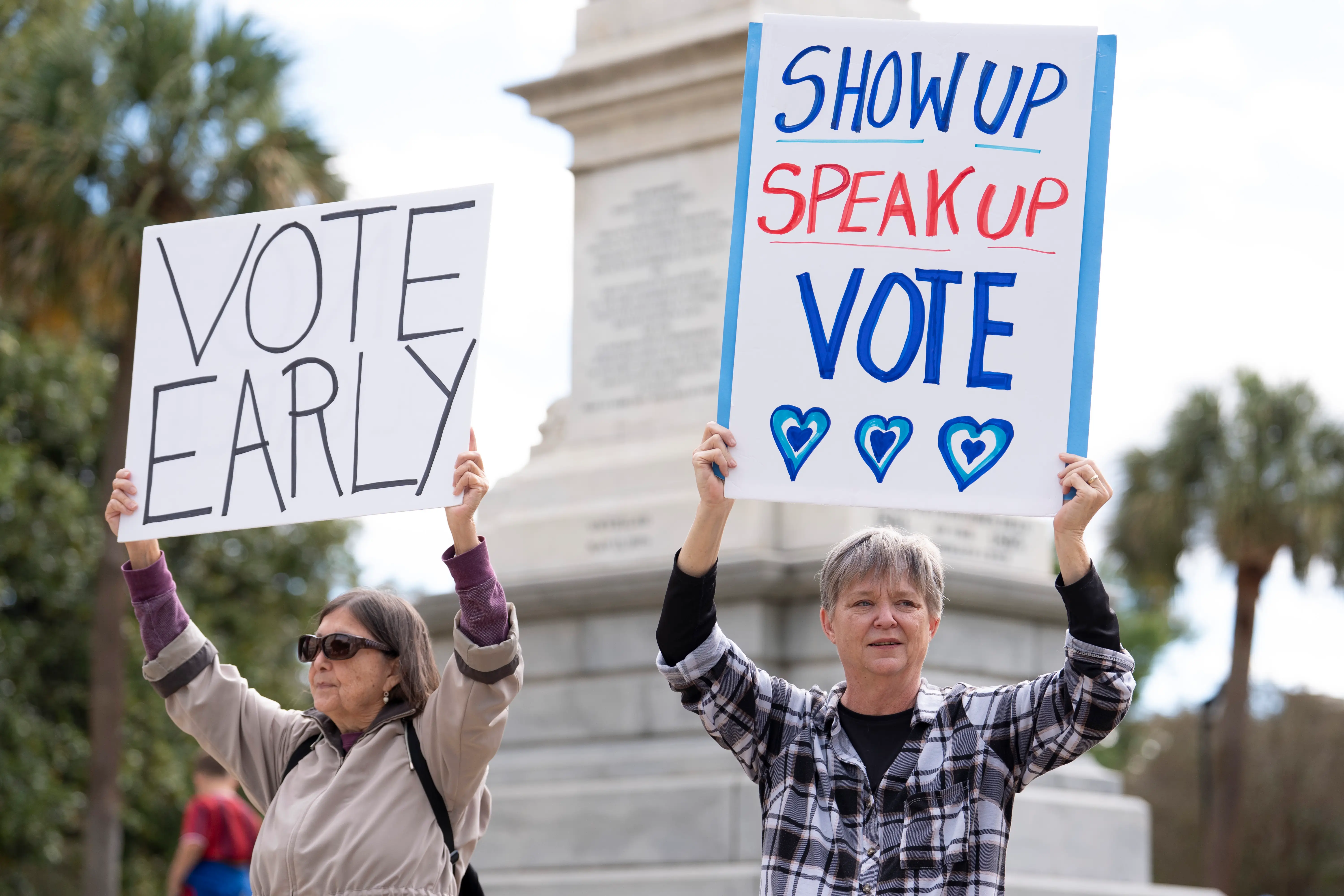 Two demonstrators hold handmade voting signs reading “VOTE EARLY” and “SHOW UP SPEAK UP VOTE” near a monument.