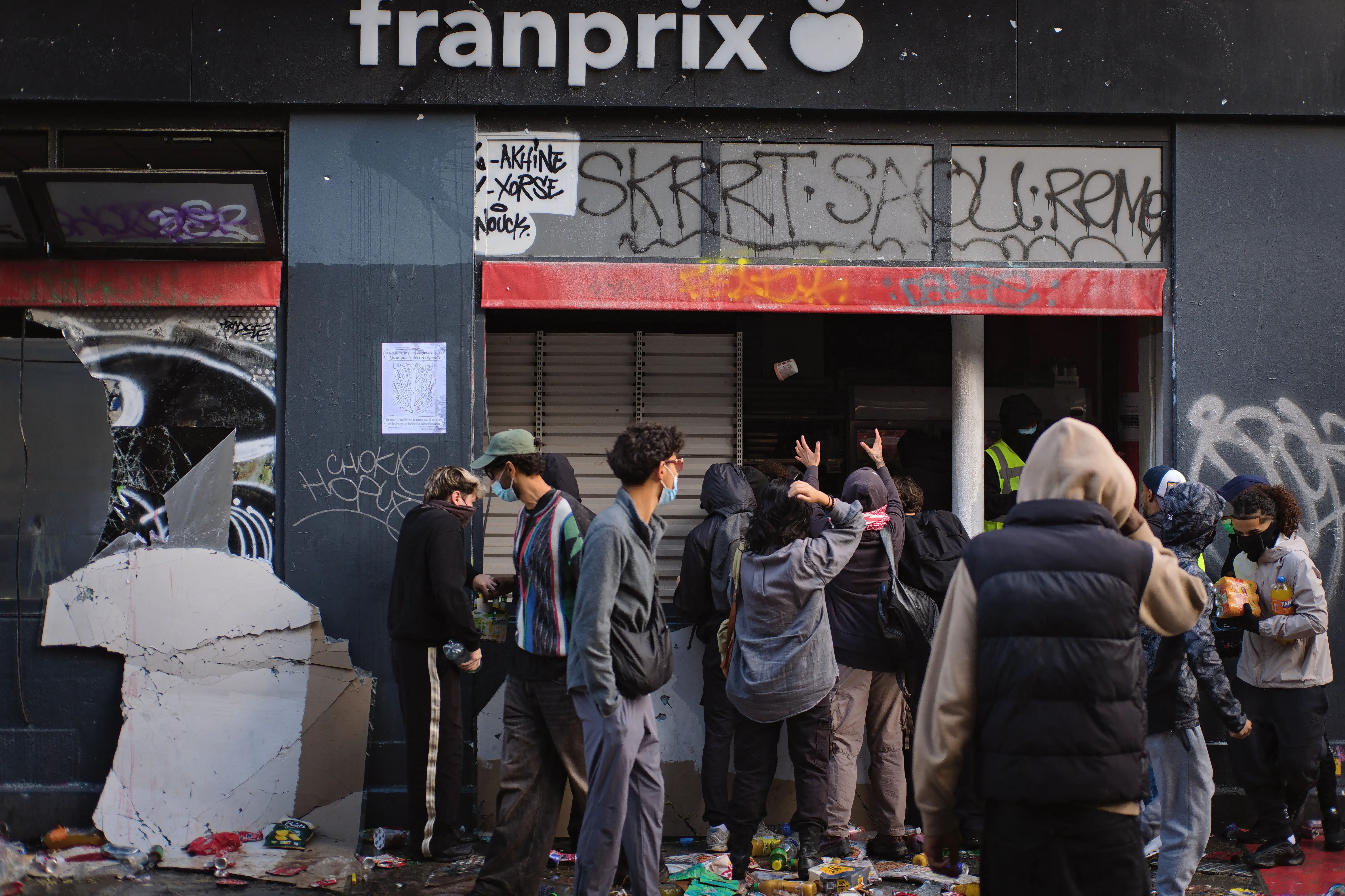 People gather outside a damaged Franprix storefront with graffiti, broken panels, and debris on the sidewalk.