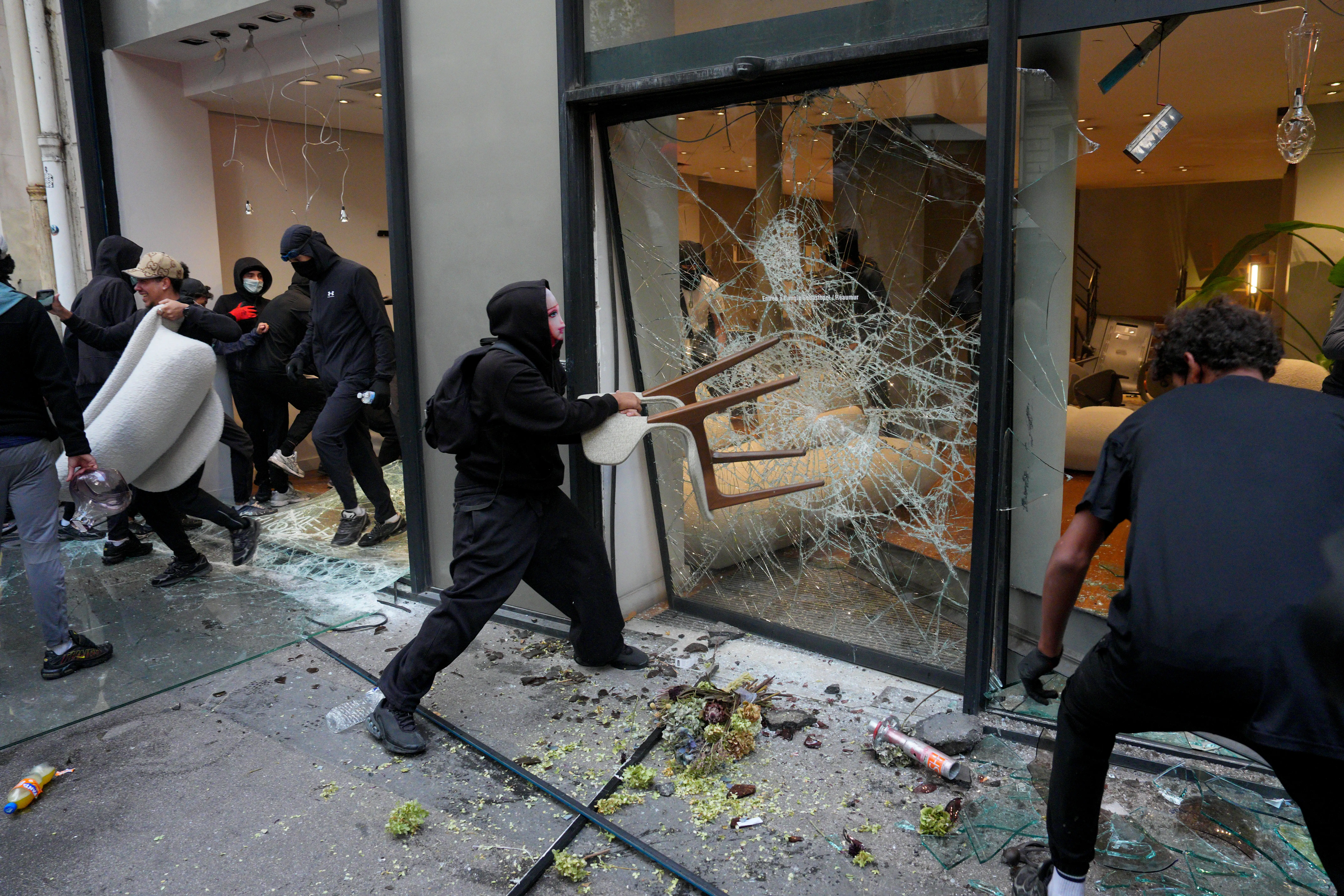 Masked people break a storefront window amid shattered glass and scattered debris on a city sidewalk.
