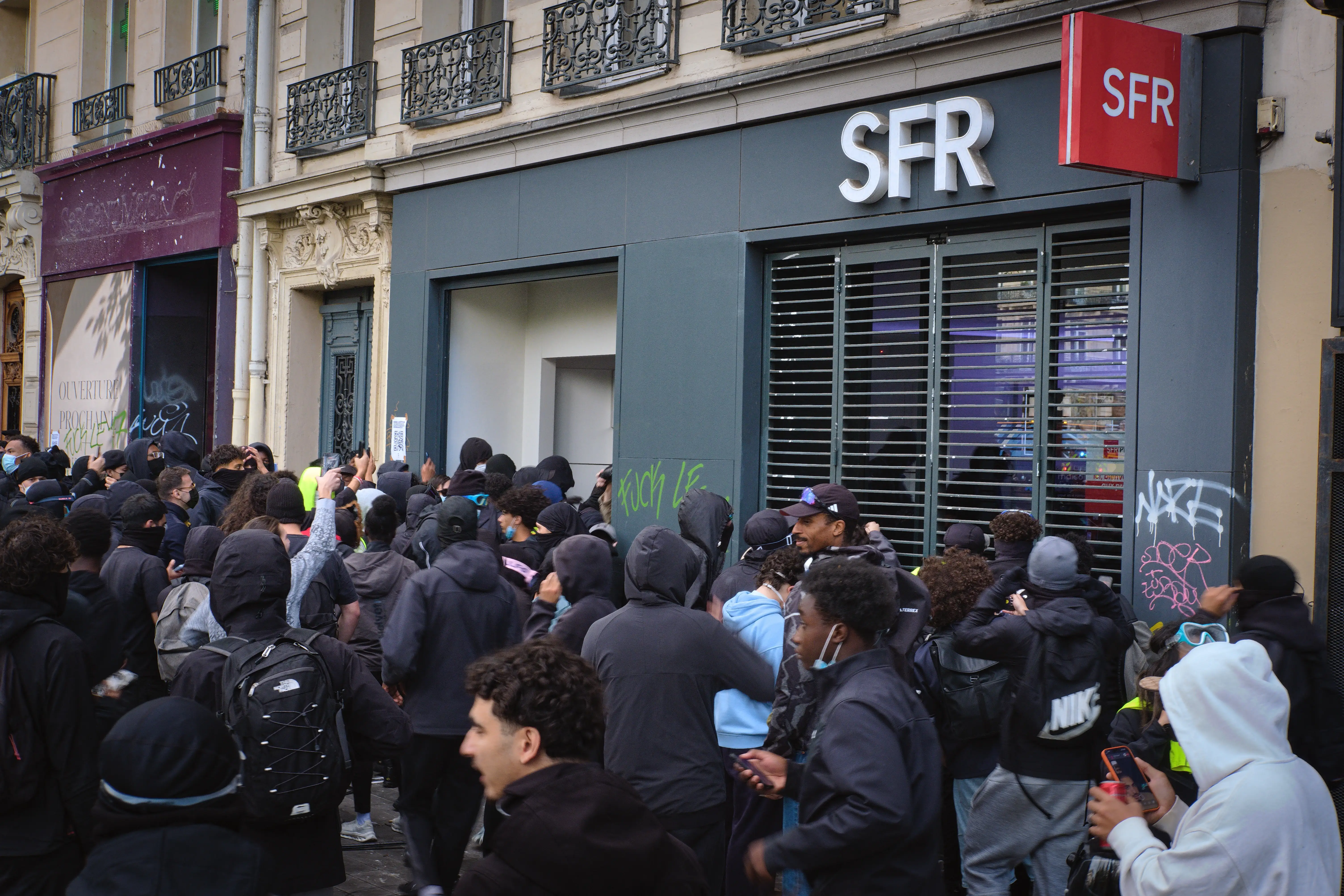 Crowd of hooded people gathers outside an SFR storefront with phones raised on an urban street.