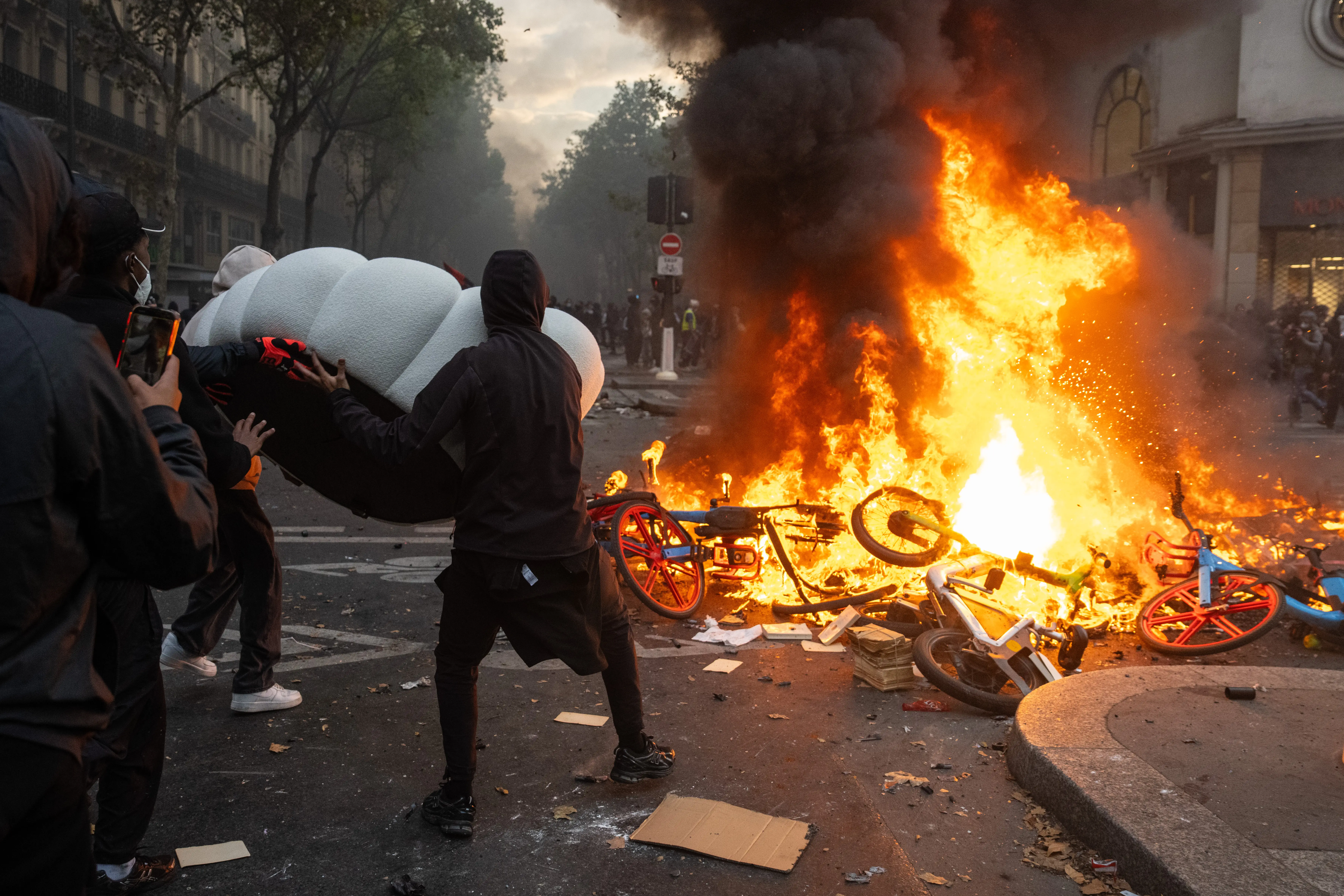 People stand near a large street fire burning bicycles and debris as thick black smoke rises in an urban area.
