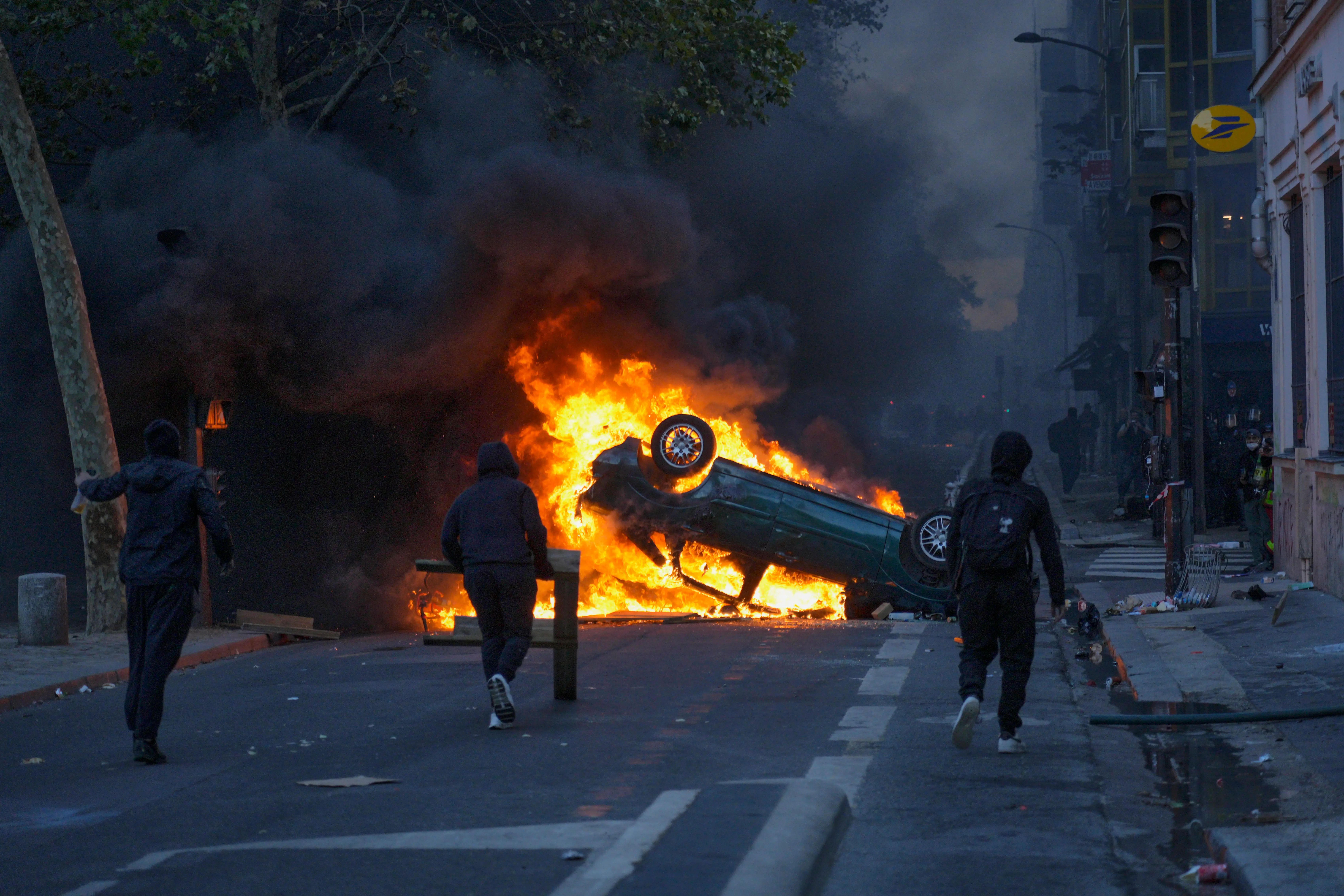 An overturned car burns in a city street as thick smoke rises and several people move nearby.