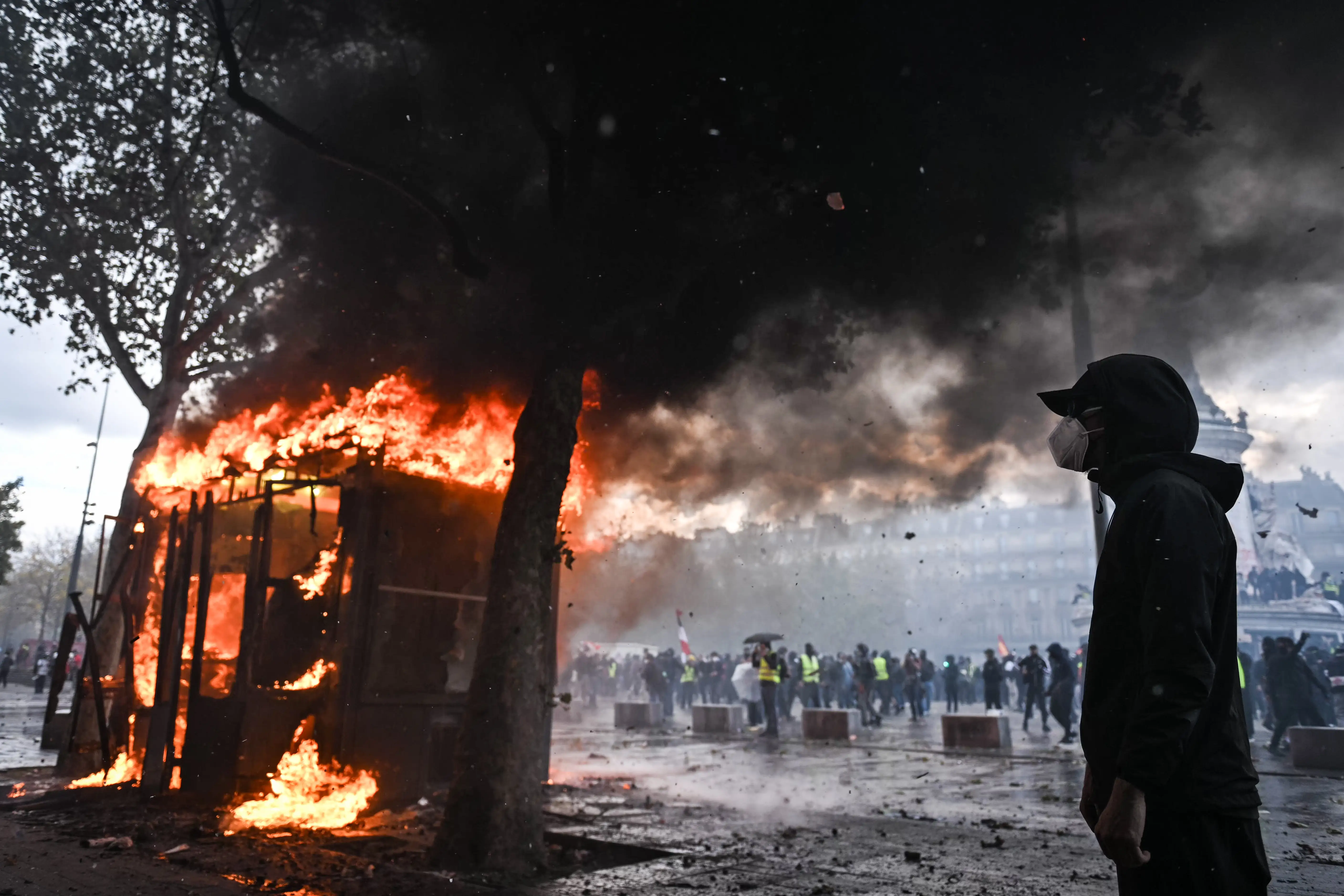 A hooded person stands near a burning street structure as smoke rises over a crowd in an urban area.