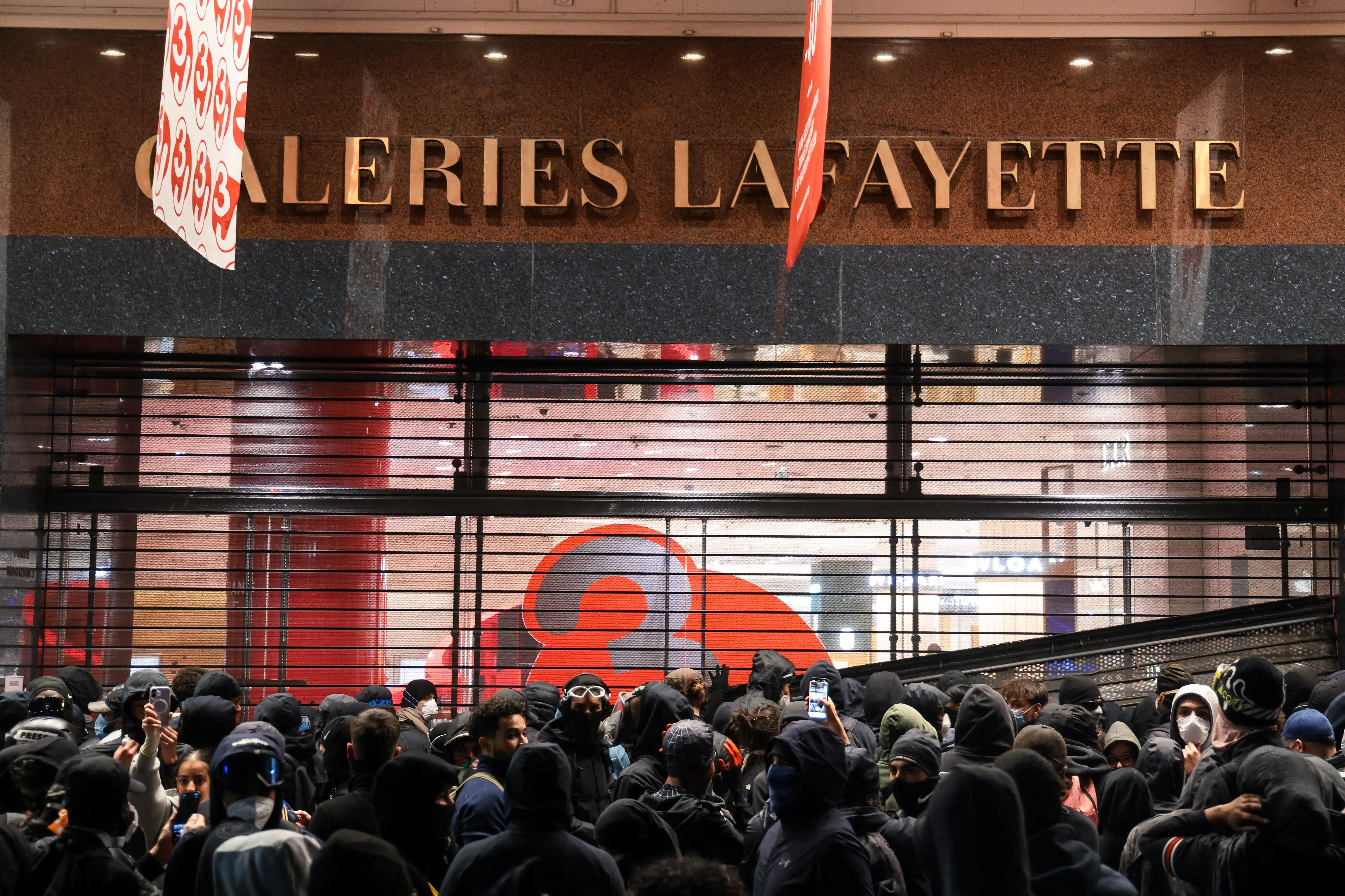 Crowd gathers outside a Galeries Lafayette storefront with a metal security grille partly covering the entrance.