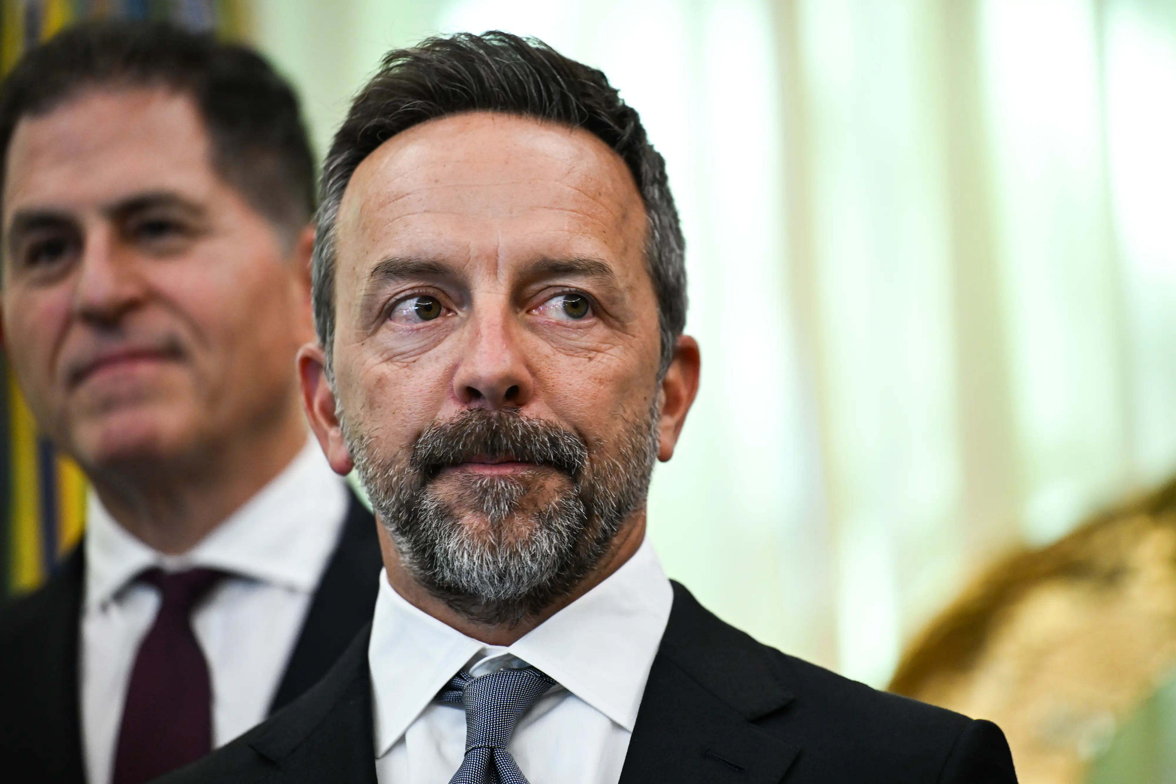 Brad Gerstner in a dark suit and tie stands in focus beside another suited person indoors.
