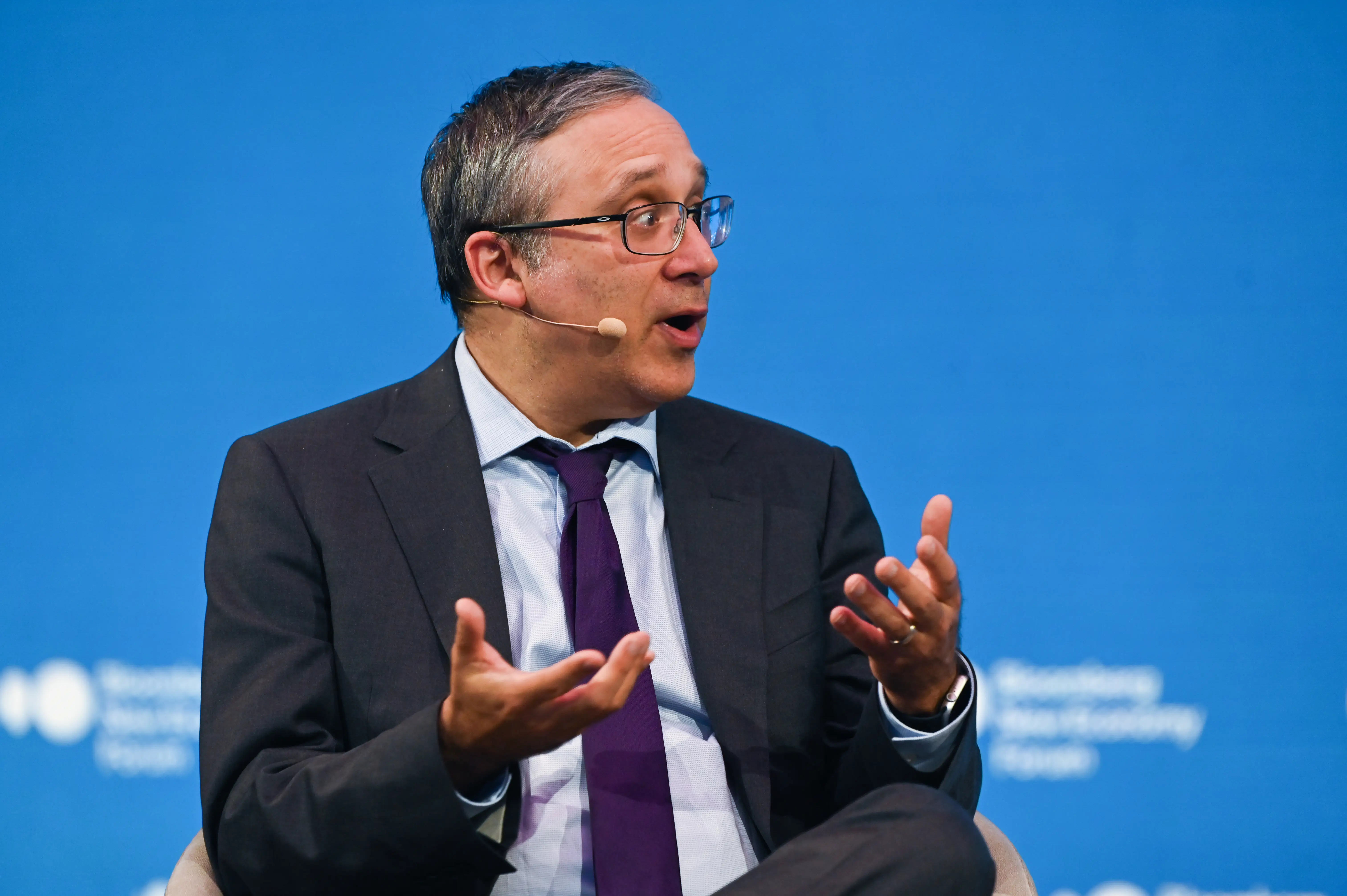 A suited speaker wearing a headset microphone gestures during a panel against a blue conference backdrop.