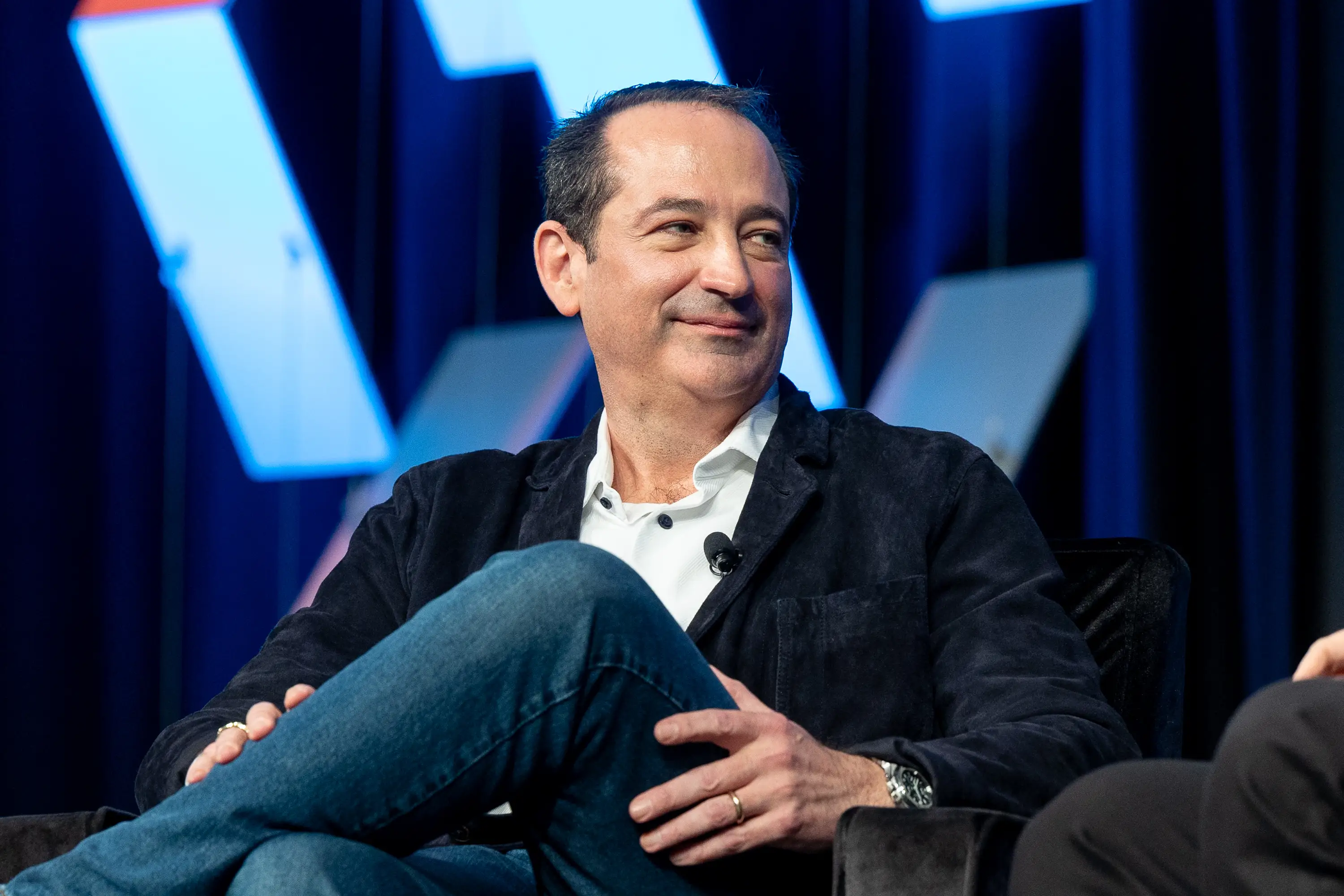 Seated speaker in a dark blazer and jeans smiles on a blue-lit conference stage.