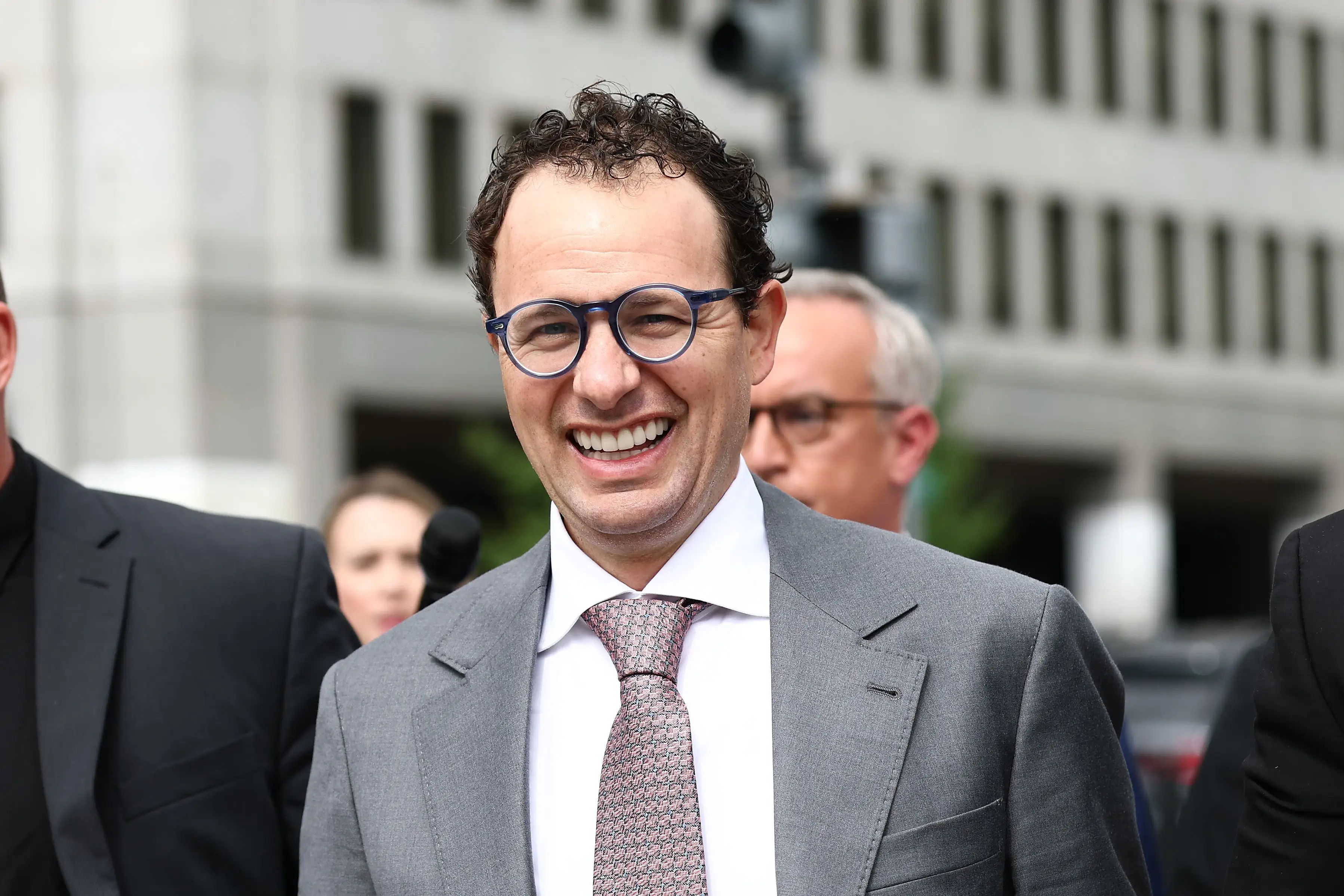 Anthropic CEO Dario Amodei smiles while walking with suited security personnel outside a city building.