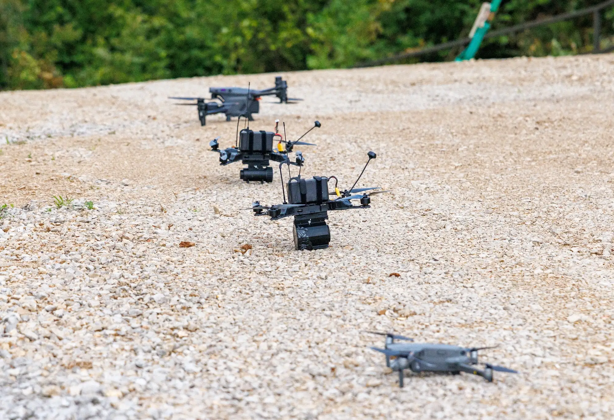 Several quadcopter drones rest in a line on a gravel path with trees in the background.
