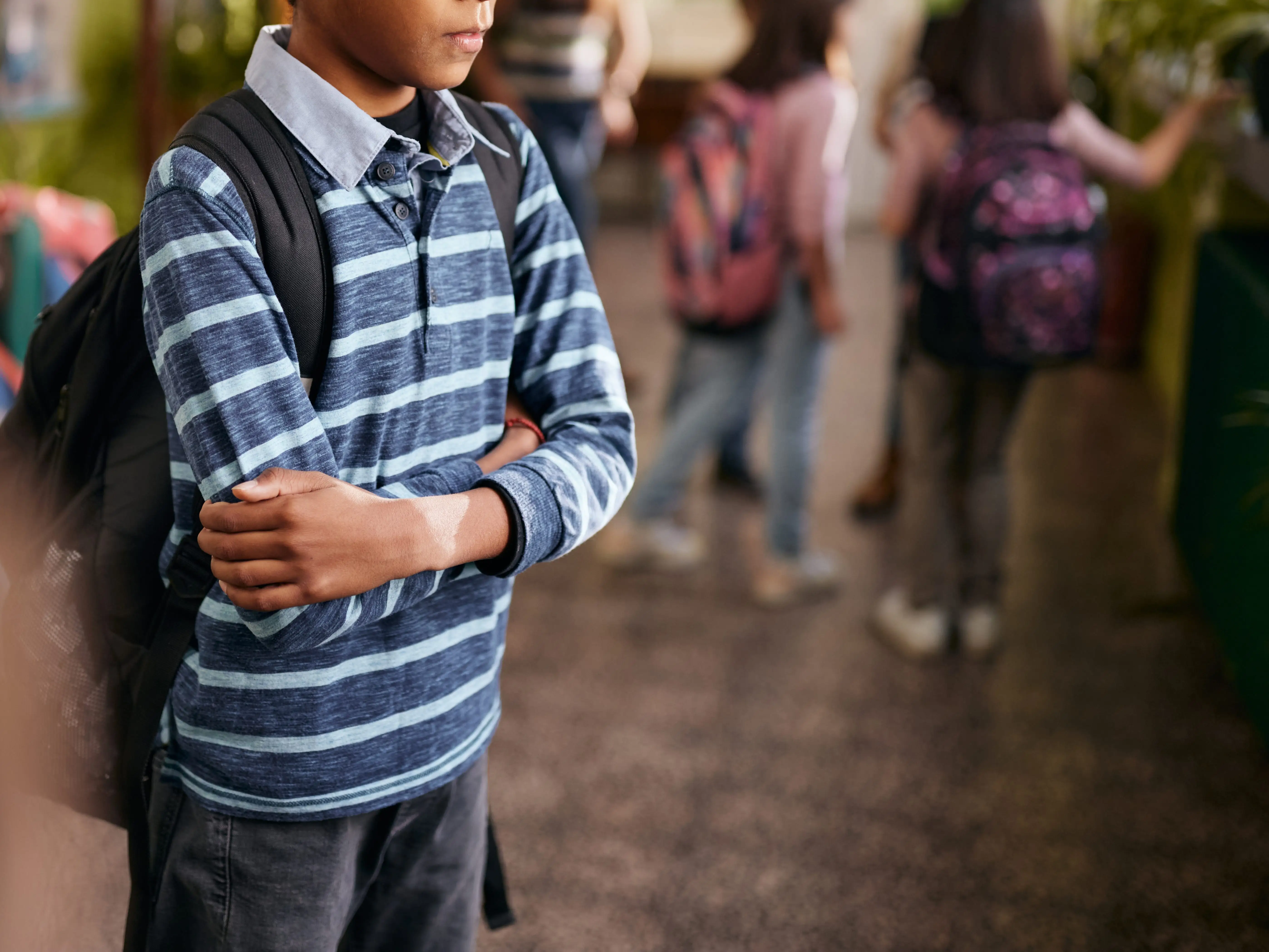 A student with a backpack stands arms crossed in a busy school hallway with classmates blurred behind.
