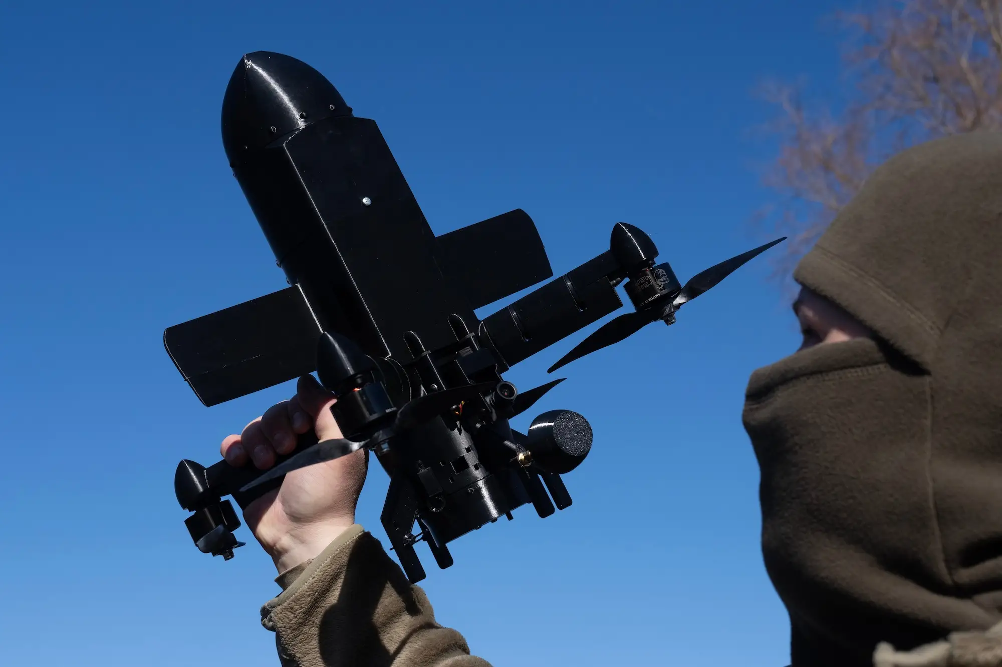 A masked person holds a black fixed-wing drone against a clear blue sky.