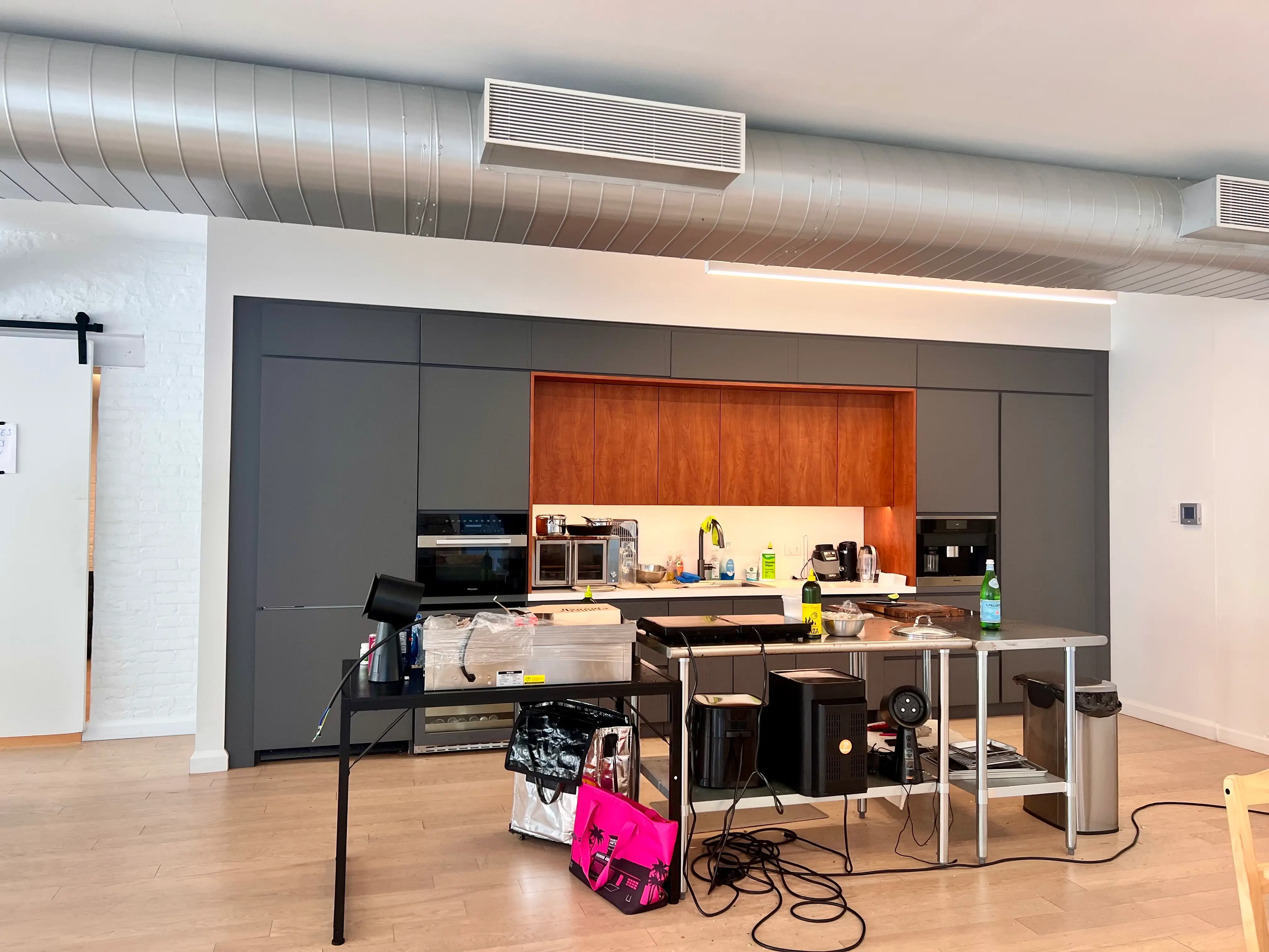 Modern open kitchen with gray cabinetry, wood accents, equipment tables, appliances, cables, and exposed ceiling ductwork.