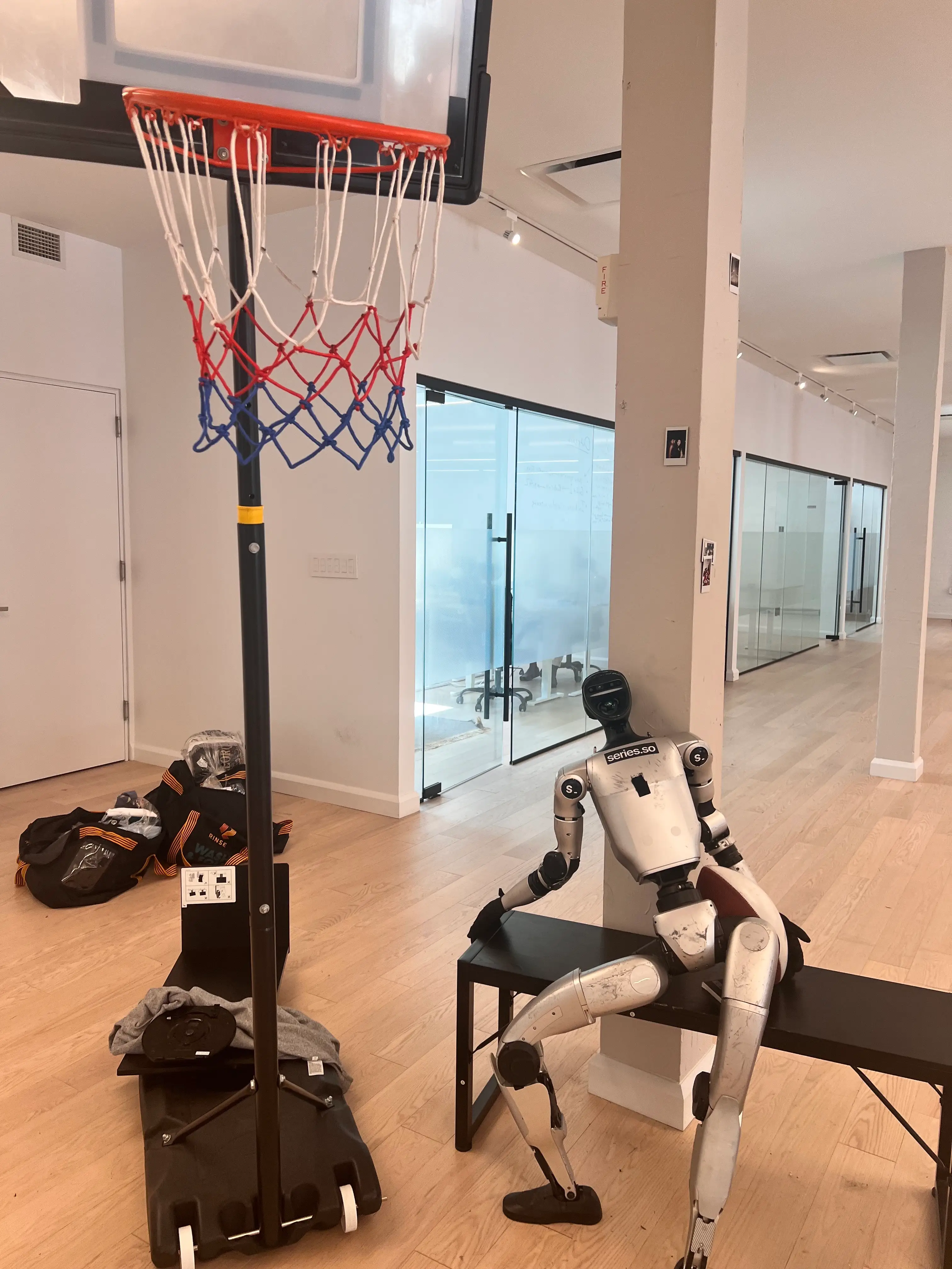 Humanoid robot sits on a bench beside a portable basketball hoop in a modern office hallway.